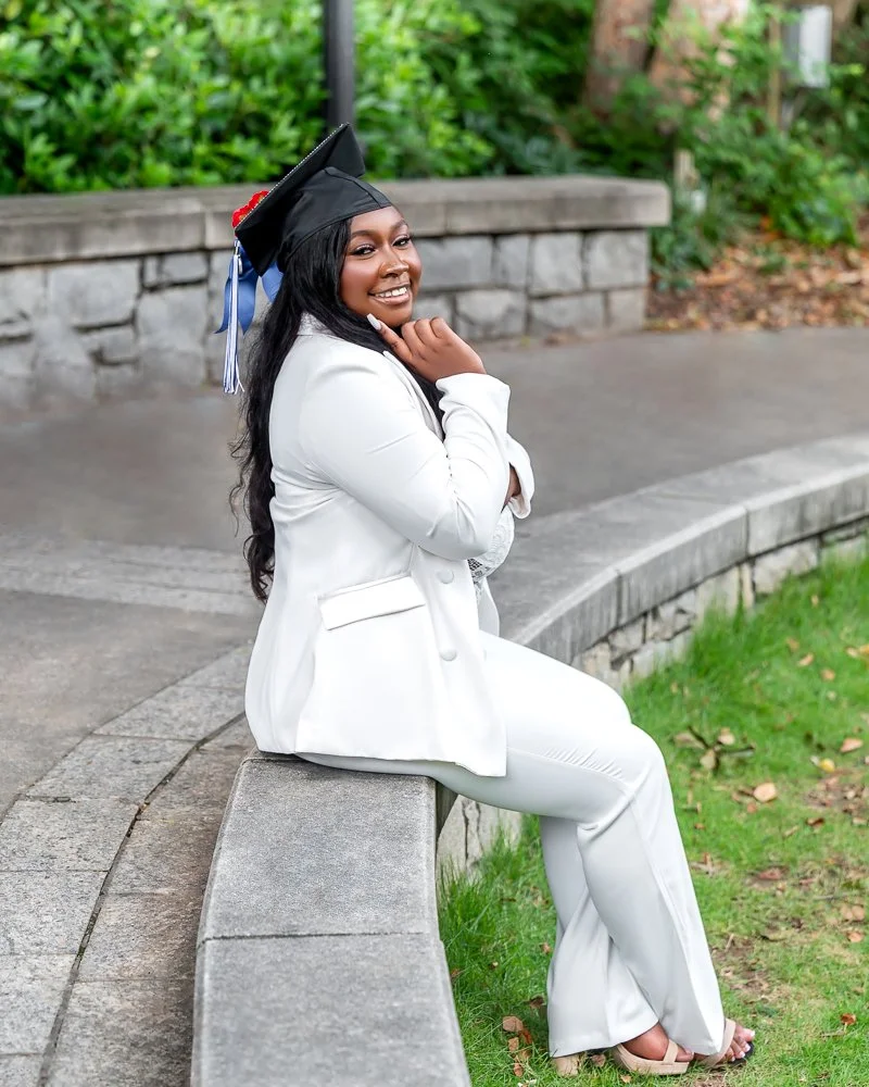A young woman in a white suit and white dress shoes sitting on a curved stone bench outdoors, wearing a black graduation cap with a blue tassel, smiling and posing with her hand near her chin.