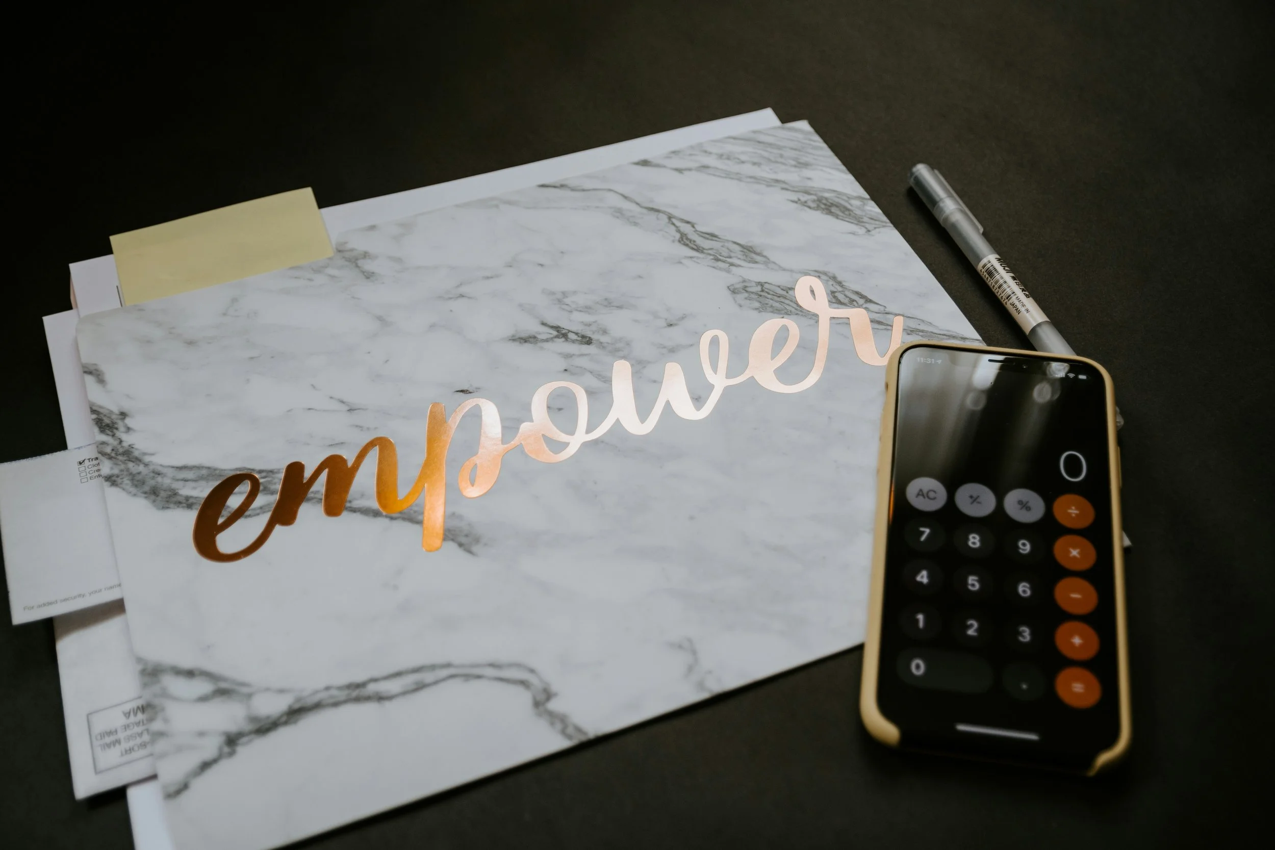 Work desk with a stack of papers, a yellow sticky note, a silver pen, a smartphone displaying a calculator app, and a sign that says "empower" on a marble-patterned paper, all on a dark surface.