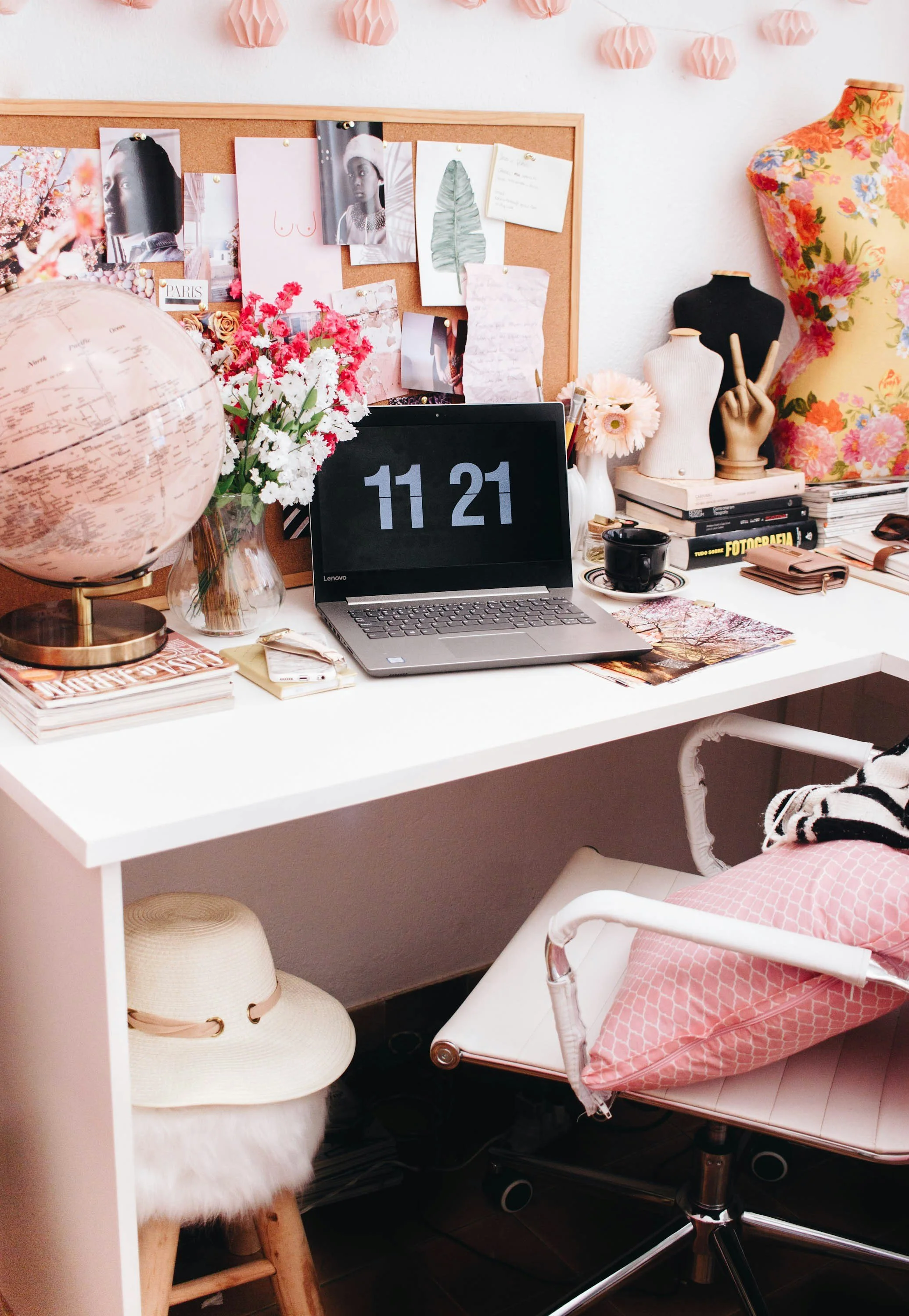 A stylish workspace with a white desk, black laptop showing the time 11:21, a globe, a bouquet of pink and white flowers, a black coffee mug, stacked books, and a pink patterned pillow on a white chair with a pink blanket. Behind are a corkboard with photos and notes, floral mannequins, and decorative pink paper lanterns on the wall.