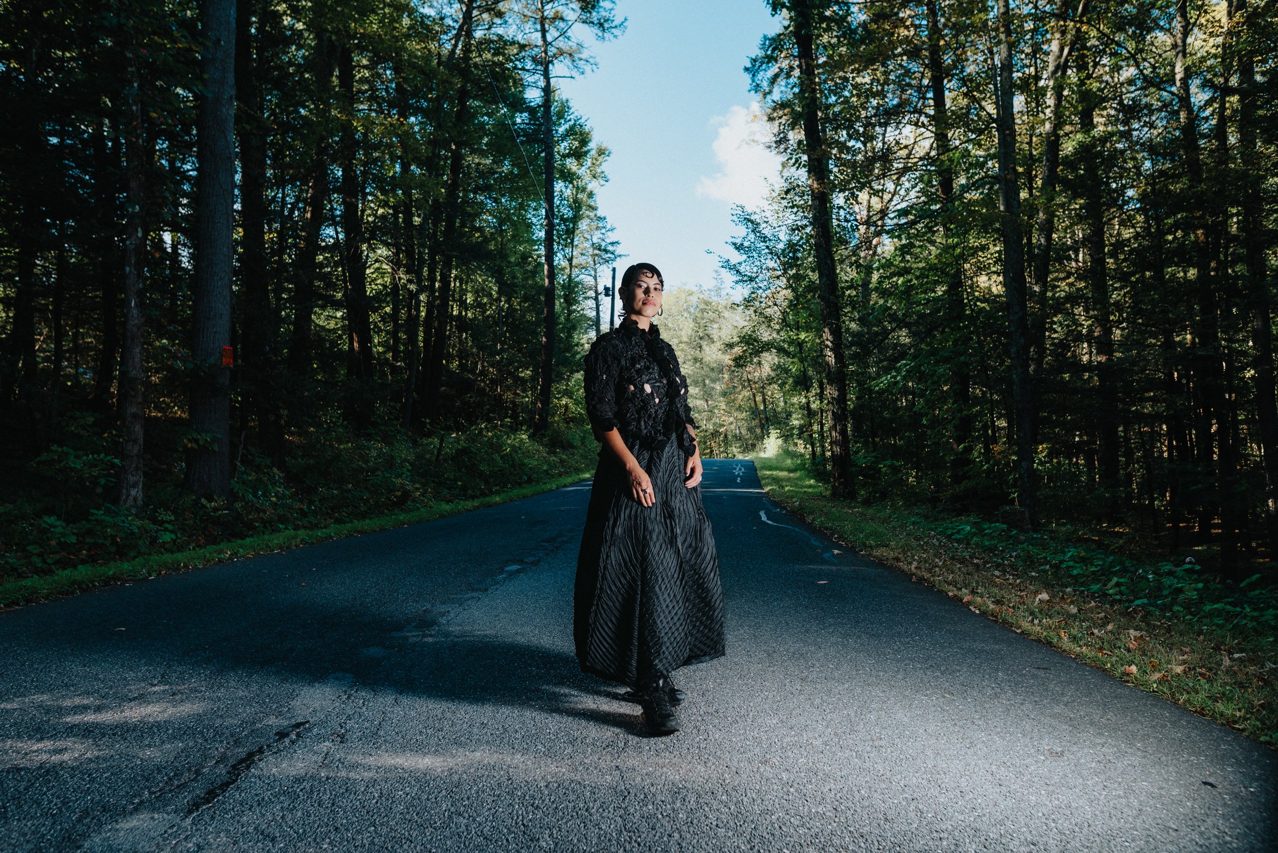 A woman in a black lace top and flowing skirt standing on a forest road with tall trees on both sides under a blue sky.