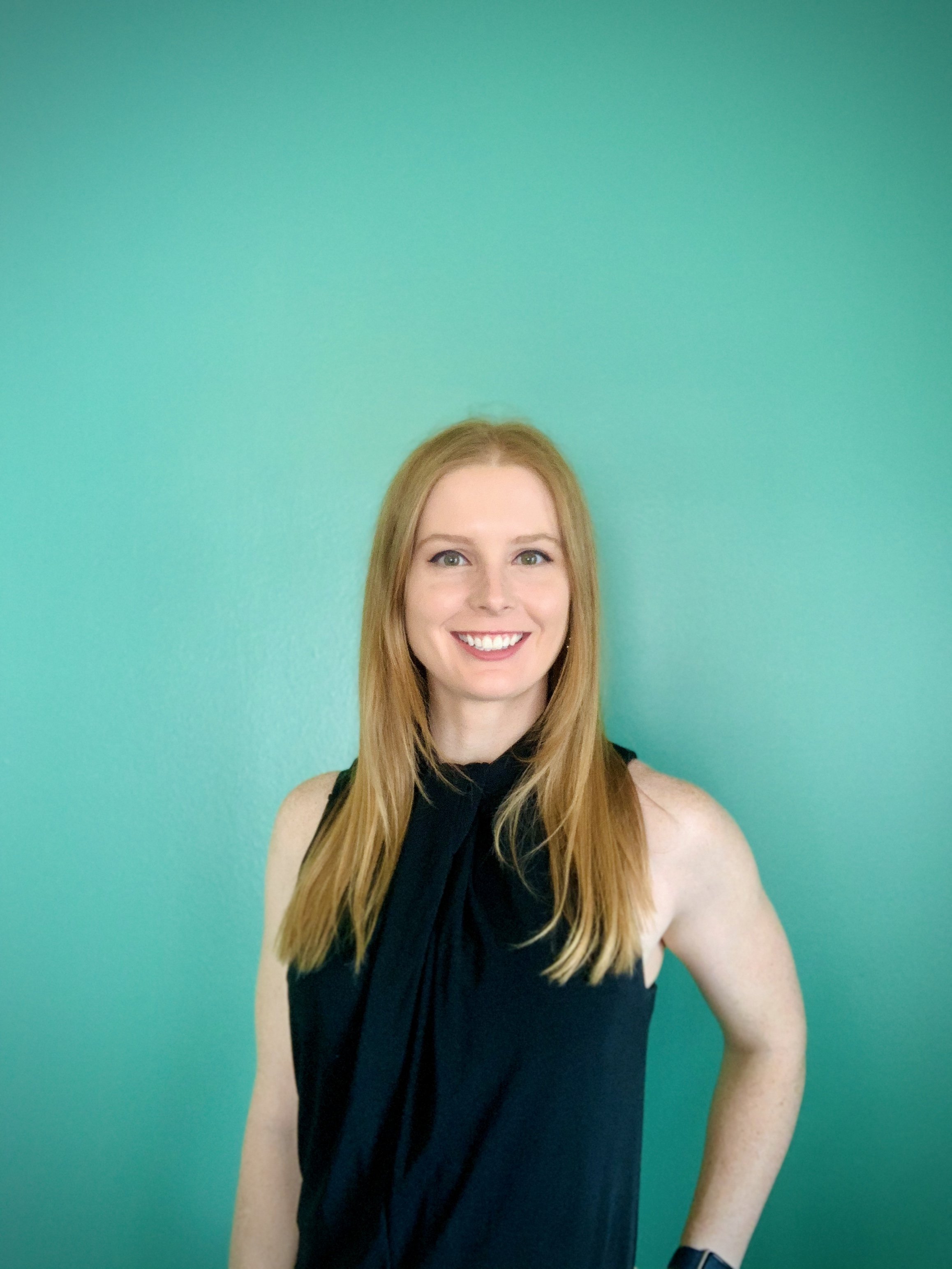 A woman with long red hair and a black sleeveless top smiling in front of a teal wall.