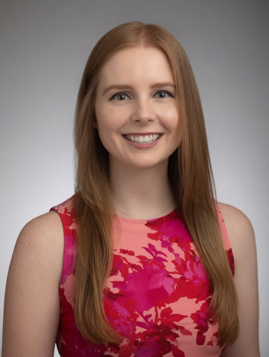A young woman with long red hair smiling, wearing a sleeveless pink and red floral dress against a gray background.