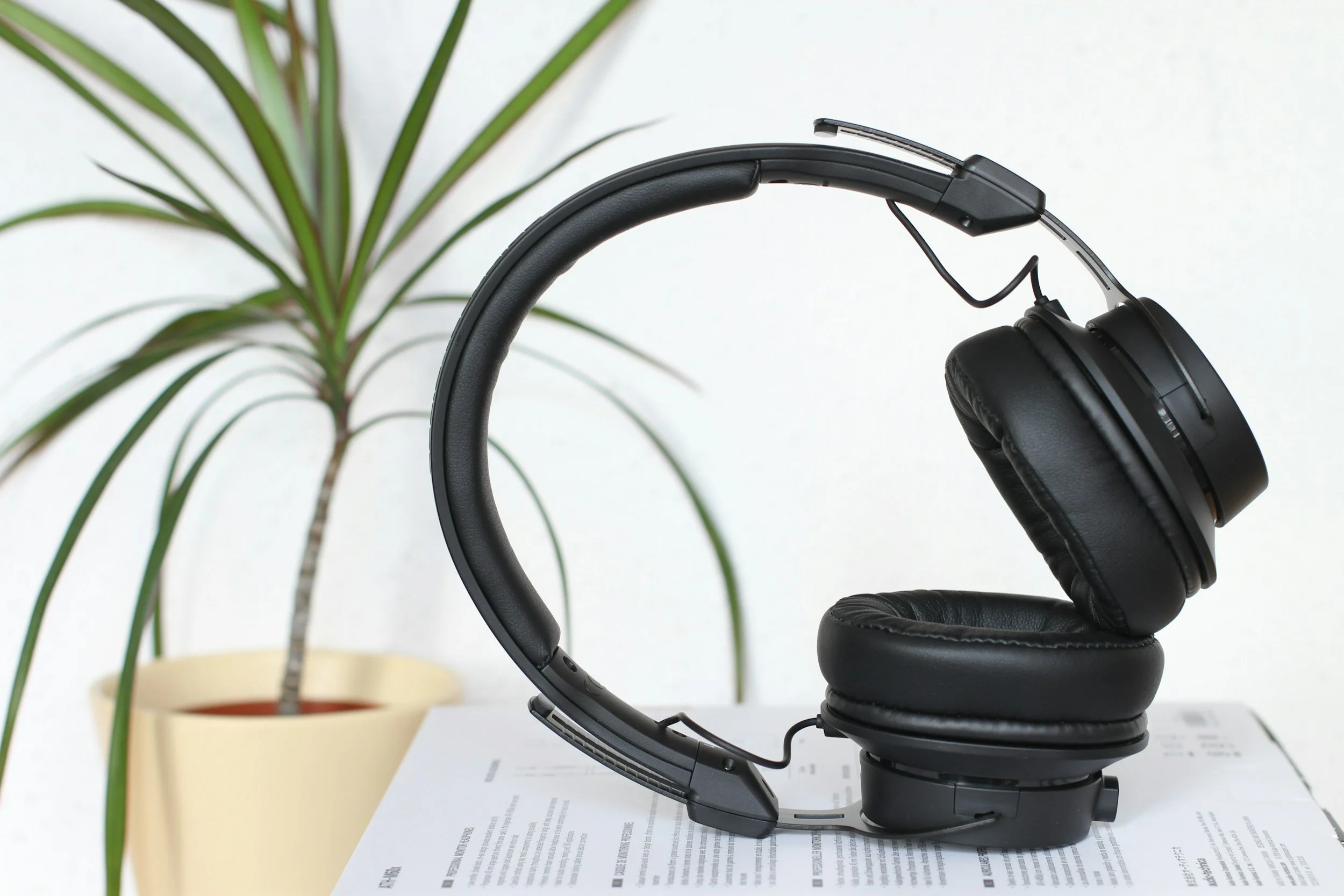 Black over-ear headphones resting on a stack of papers on a table, with a potted plant in the background.