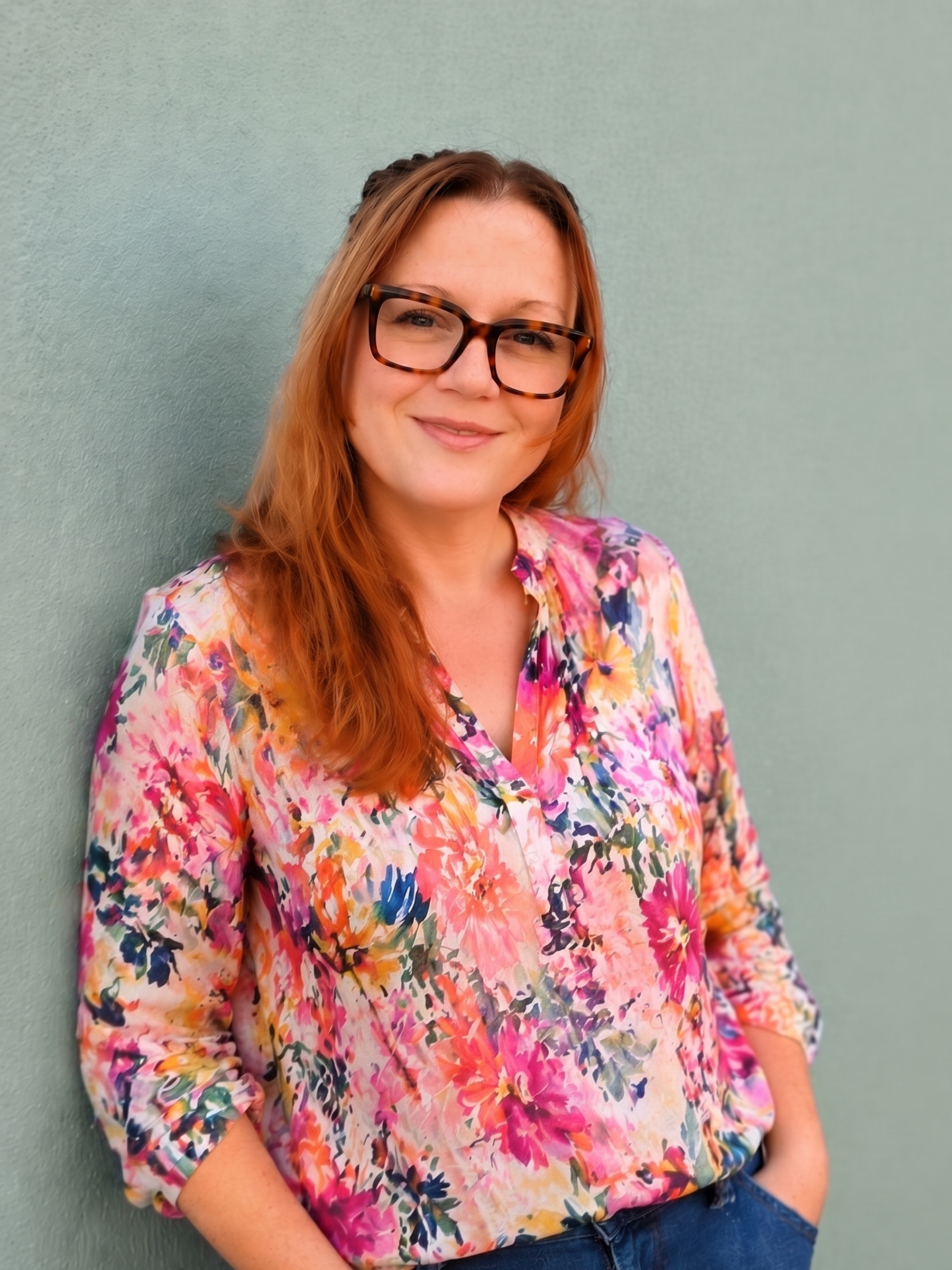 A woman with red hair and glasses wearing a colorful floral blouse, standing against a plain light green wall.