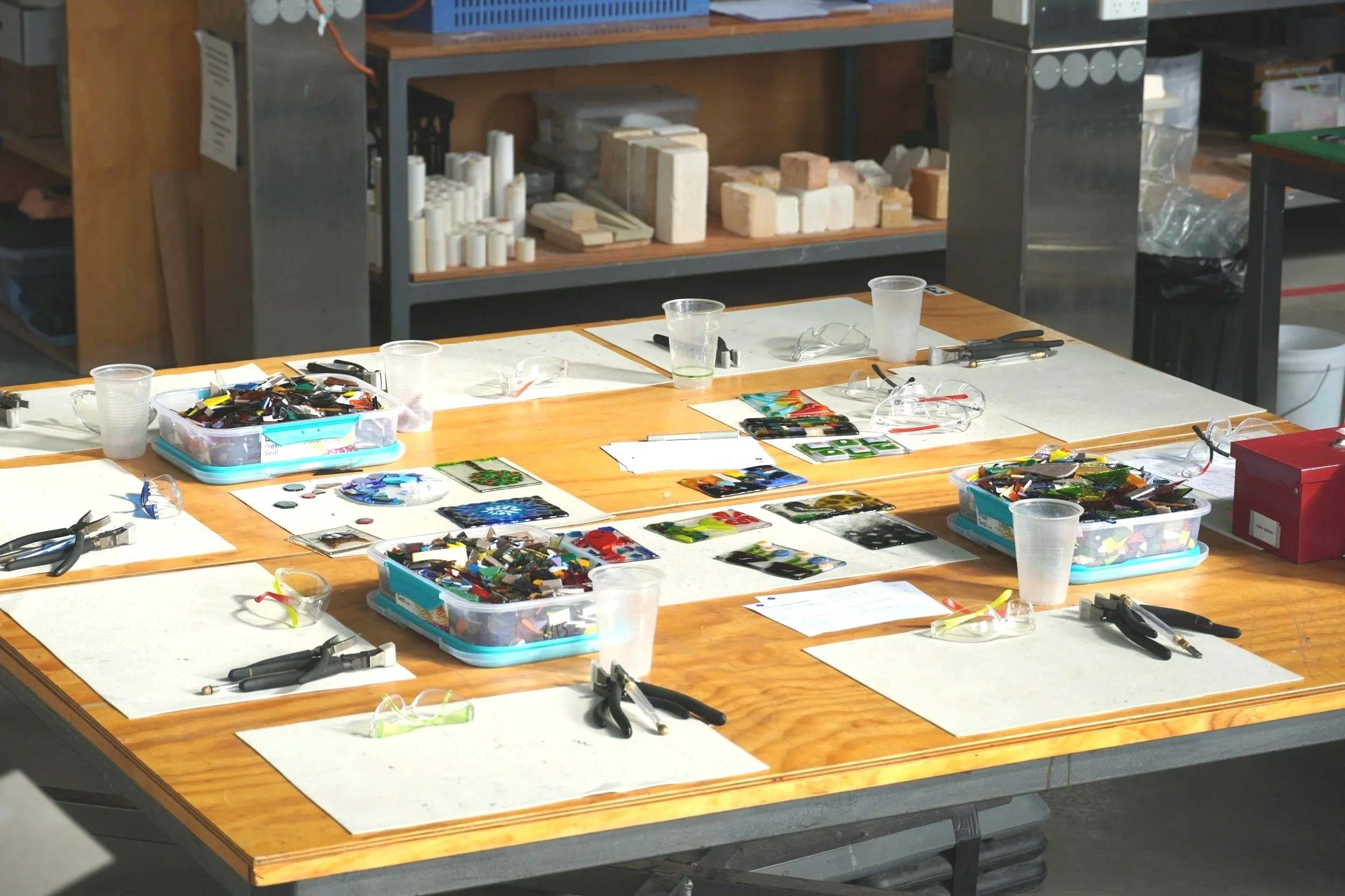 A workshop table with various artistic glass pieces, tools, safety glasses, and cups, surrounded by shelves with bricks and other materials.