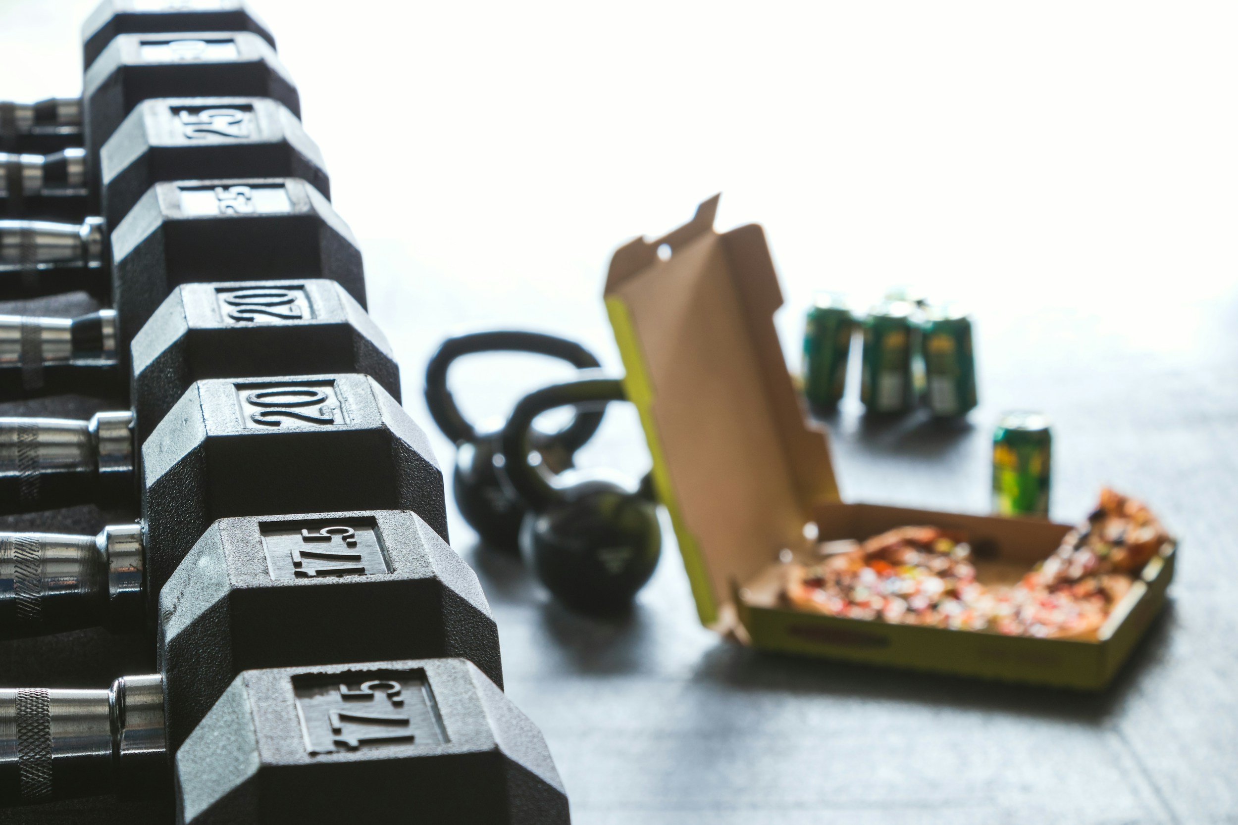 A row of black and silver adjustable dumbbells with engraved weight markings, positioned on a rack in the foreground. In the background, a box of assorted candies and chocolates in colorful wrappers, along with some small snack packages, are slightly out of focus.
