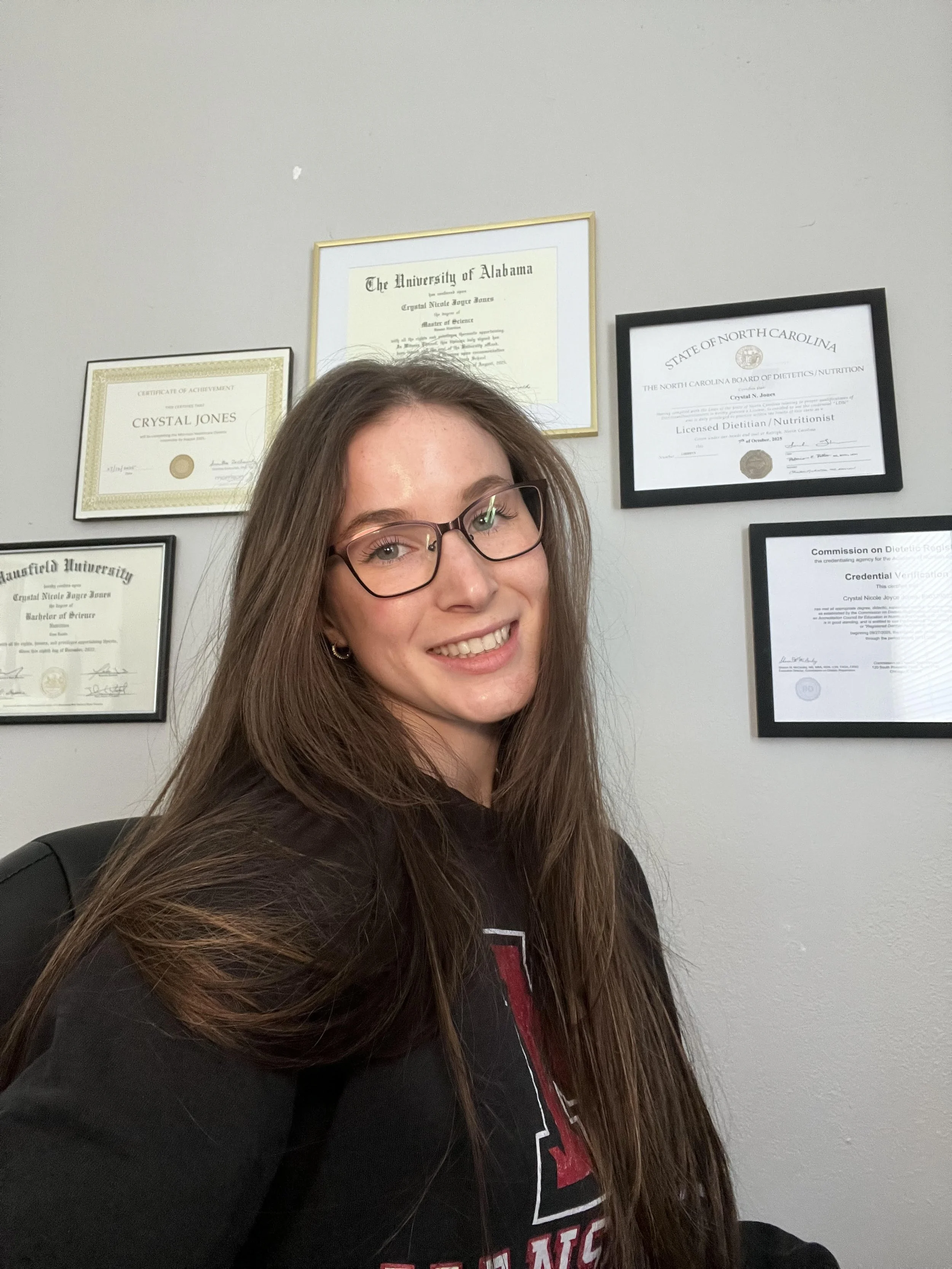 A woman with long brown hair and glasses smiling in front of a wall with framed diplomas and certificates.