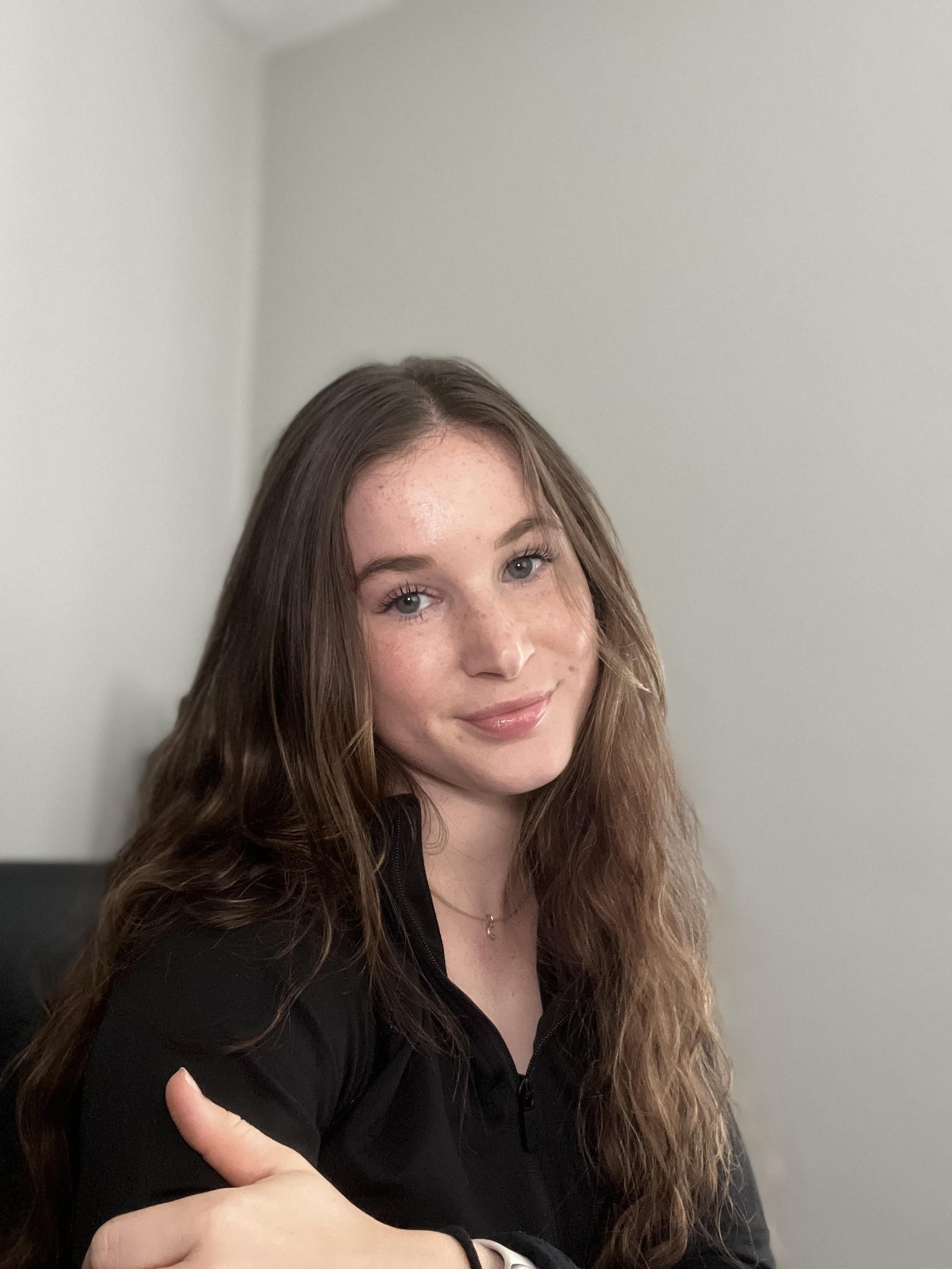 A young woman with long, wavy, brown hair smiling and crossing her arms, wearing a black top and a watch, standing against a plain, light-colored wall.