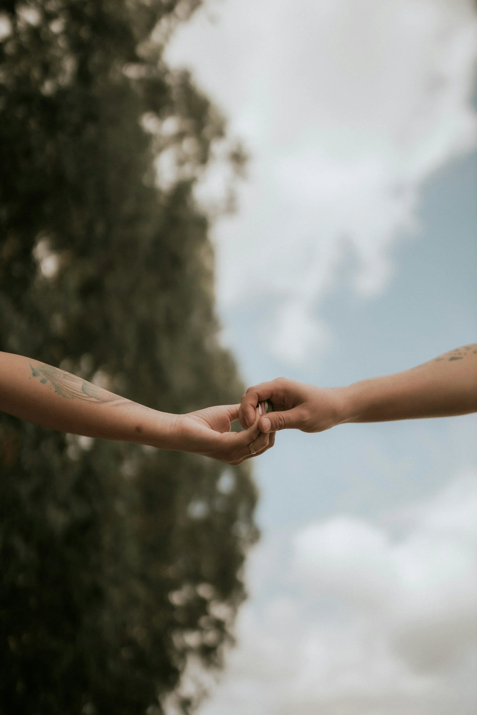 Two people are holding hands outdoors, with one person having a tattoo on their arm, against a background of cloudy sky and trees.