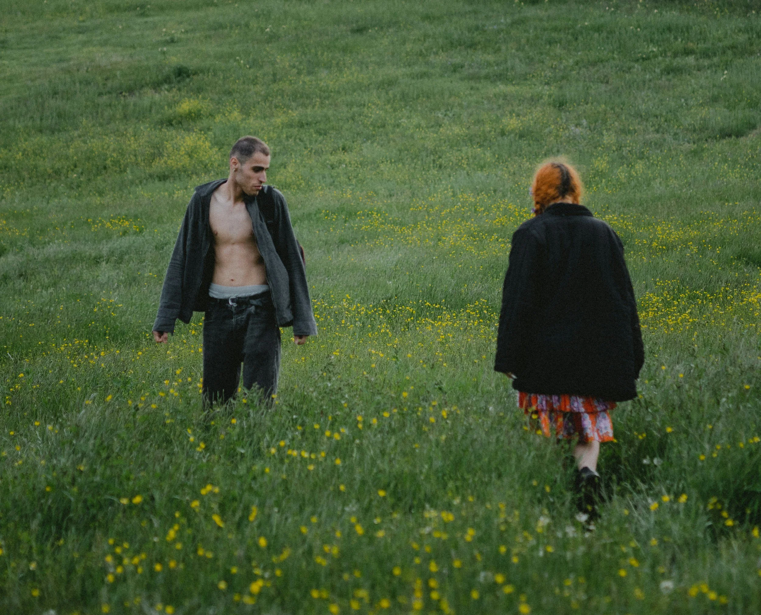 A young man with short hair and earrings, shirtless and wearing open black jacket, standing in a green field with yellow flowers, facing slightly downward. A young girl with curly hair and wearing a black jacket and a colorful dress is walking away from him.