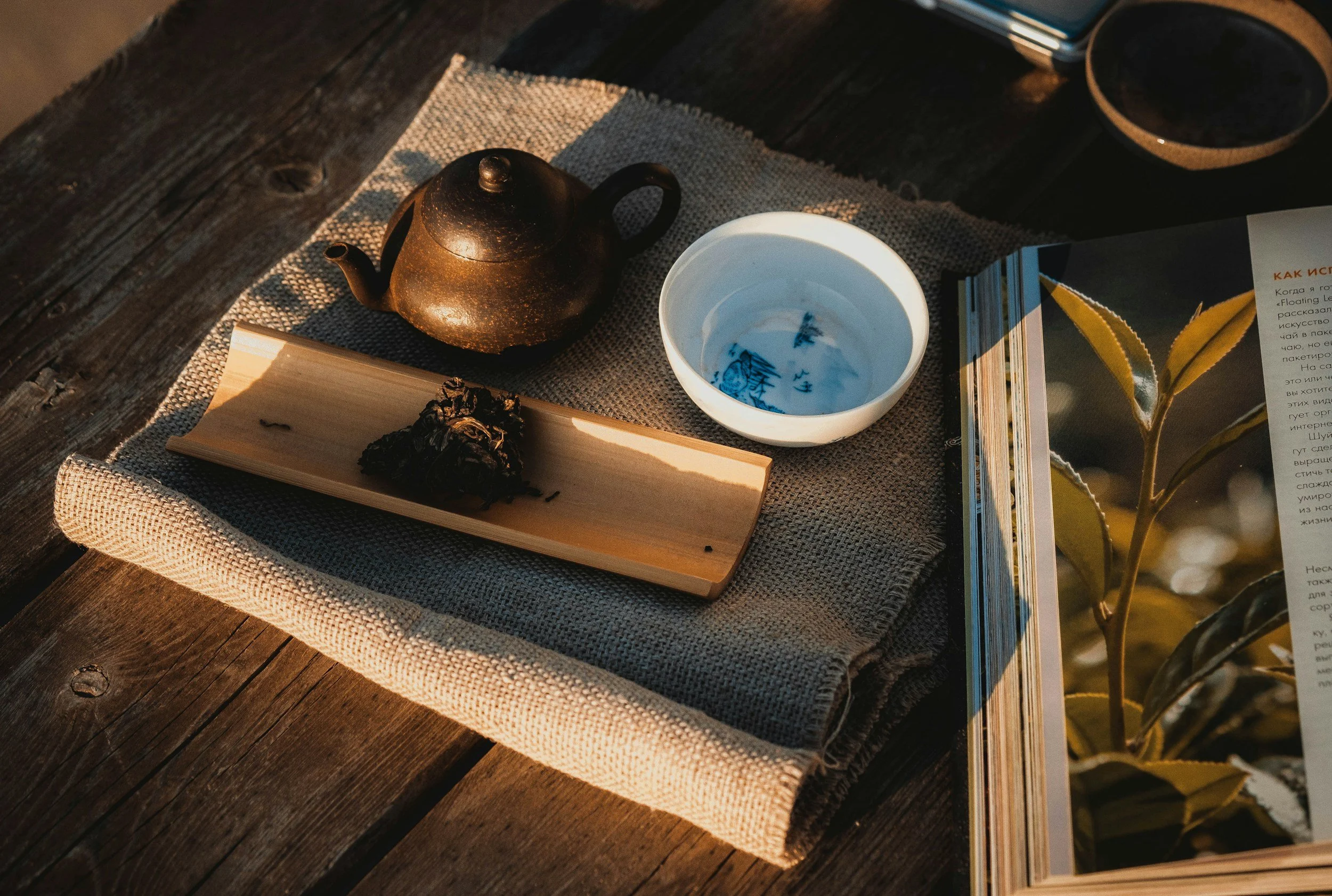 A wooden table with a beige tablecloth, featuring a black cast iron teapot, an empty white teacup with blue patterns, a small black bowl, a wooden tray with a cluster of dried herbs, a book, and some scattered papers.