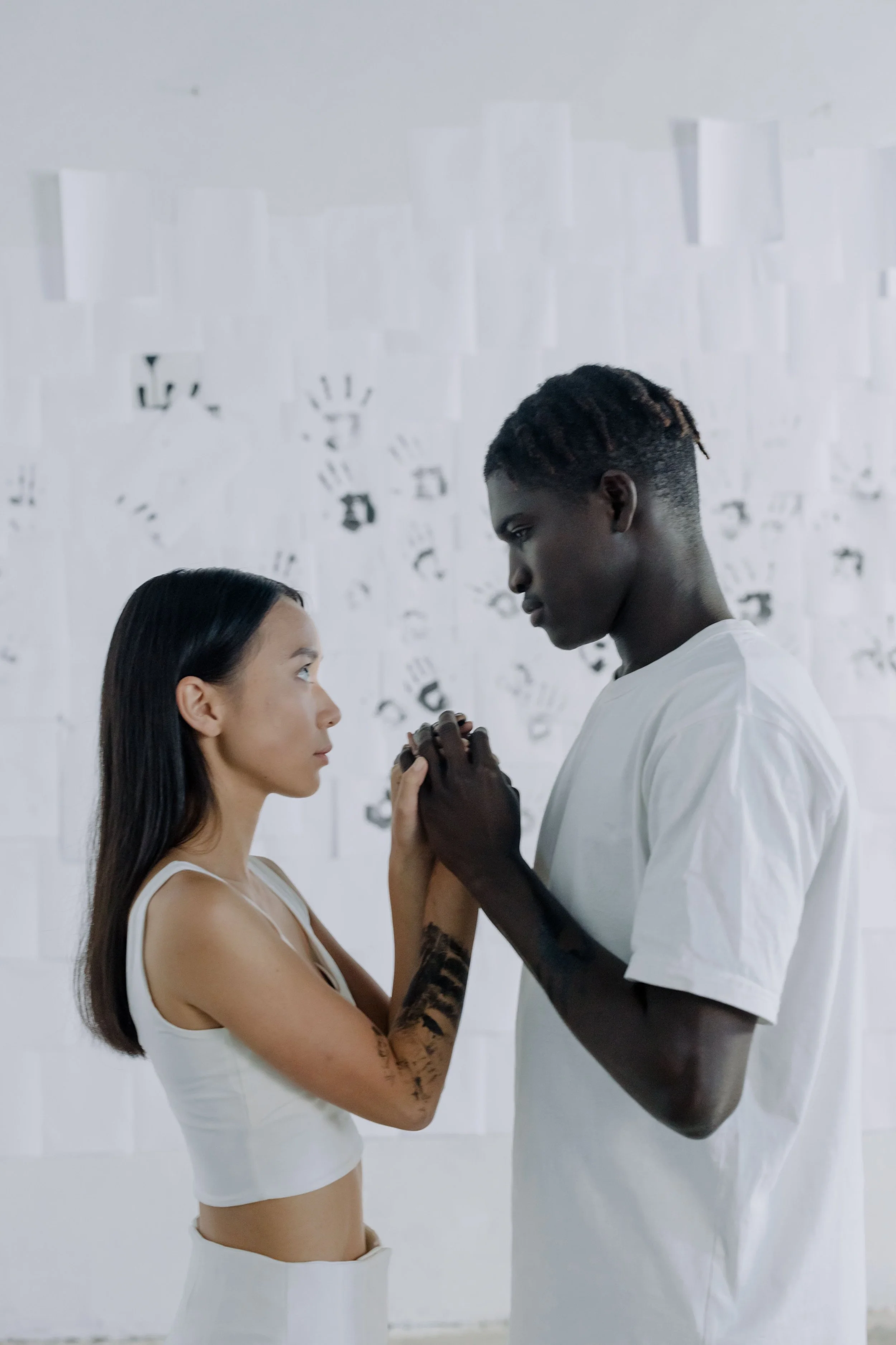 A woman with long dark hair and tattoos on her arm faces a tall man with short hair in a white shirt. They stand close, holding hands, looking into each other's eyes in front of a white wall decorated with black handprints.
