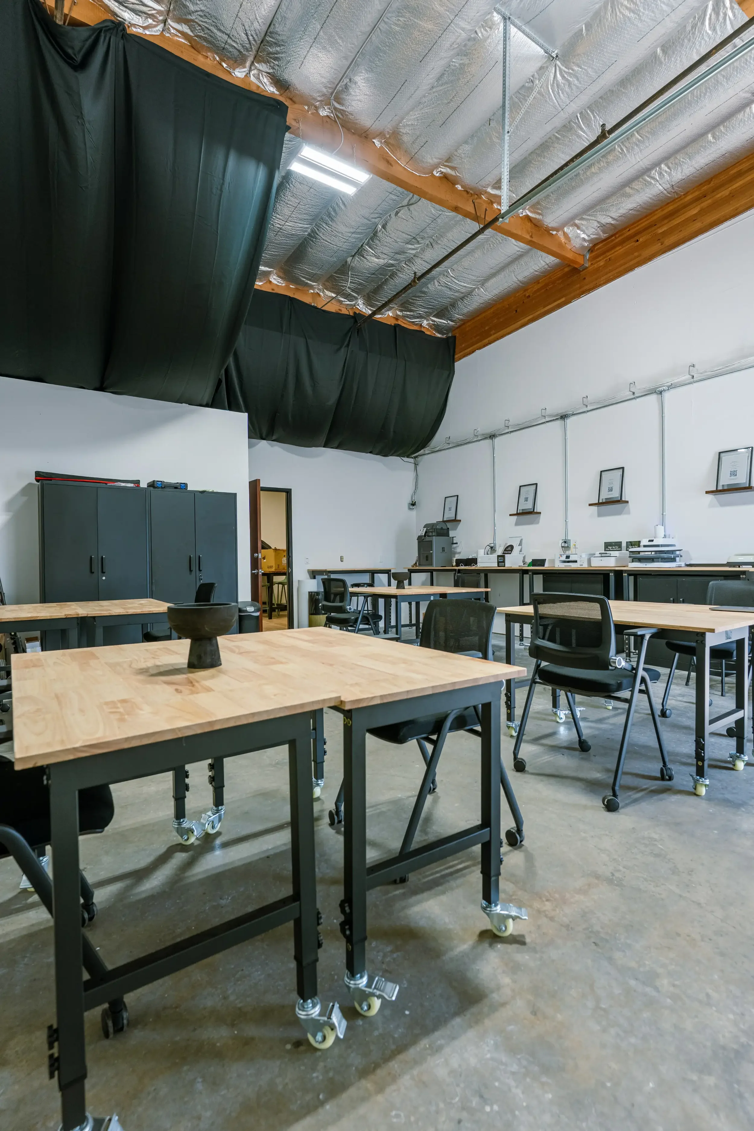 Interior of a modern gym or fitness studio with black workout tables arranged in rows, wood-paneled walls, and an industrial ceiling with exposed ductwork and large hanging lights.