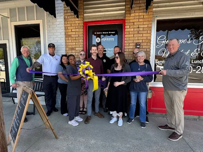 Group of people participating in a ribbon-cutting ceremony outside a storefront, with a man on the right holding scissors and purple ribbon, and others smiling around.