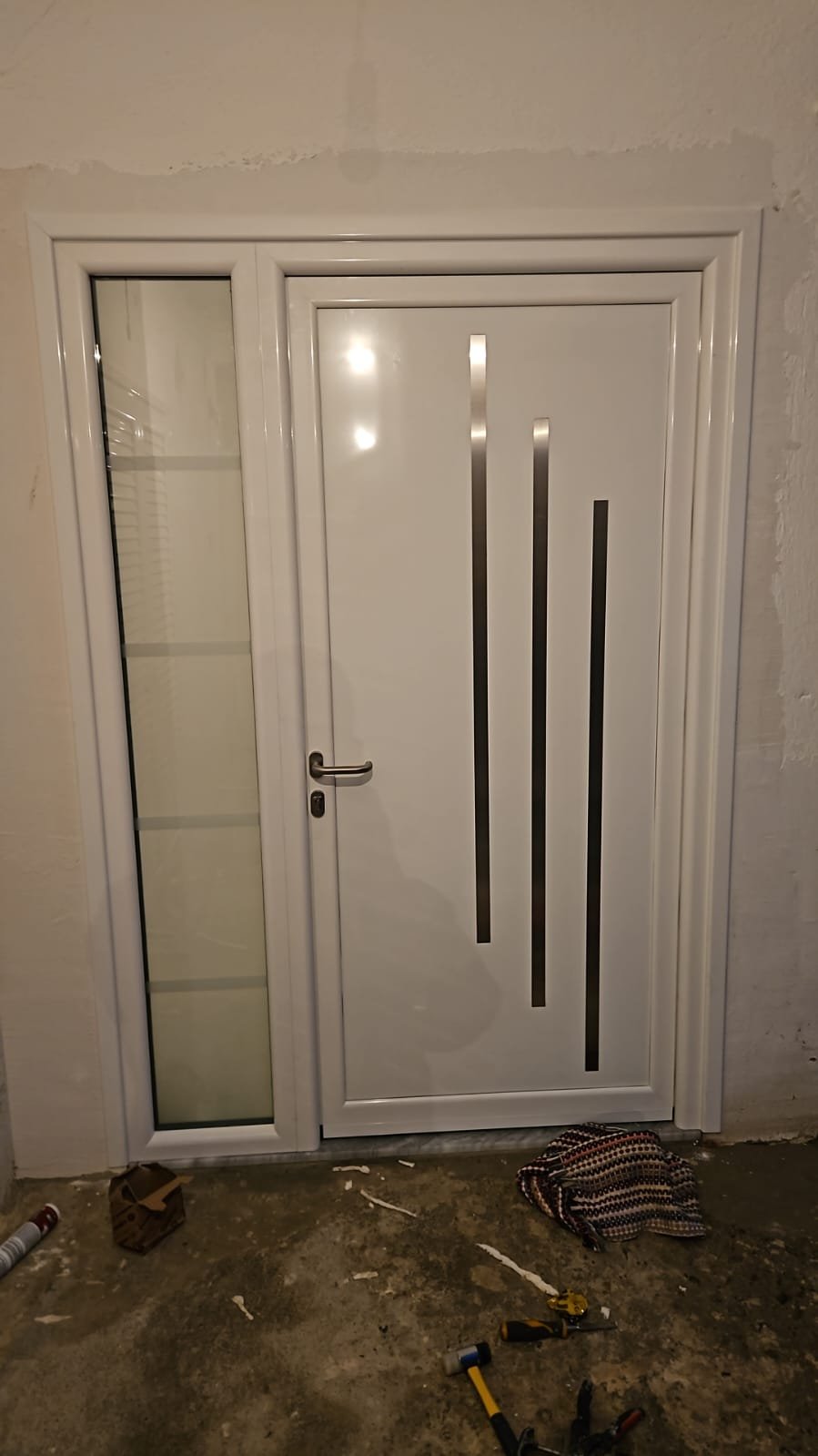 Modern white door with vertical metallic design accents, adjacent to a narrow glass window with shelves, set in a wall under construction.