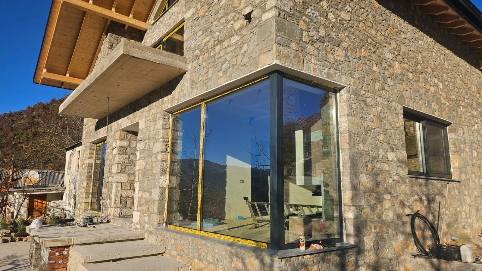 Construction site of a house with stone exterior walls, large window with black frame, and wooden balcony above. The house is unfinished, with tools and materials visible, and the sky is clear and blue.