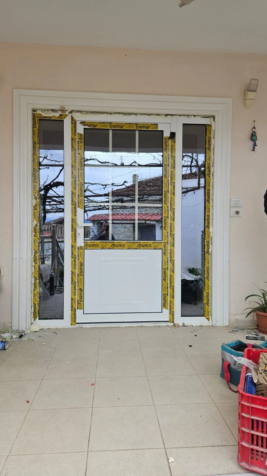 A newly installed door with side windows, still under construction, displaying protective yellow tape around the frame, with construction tools and supplies scattered on the tiled floor nearby.