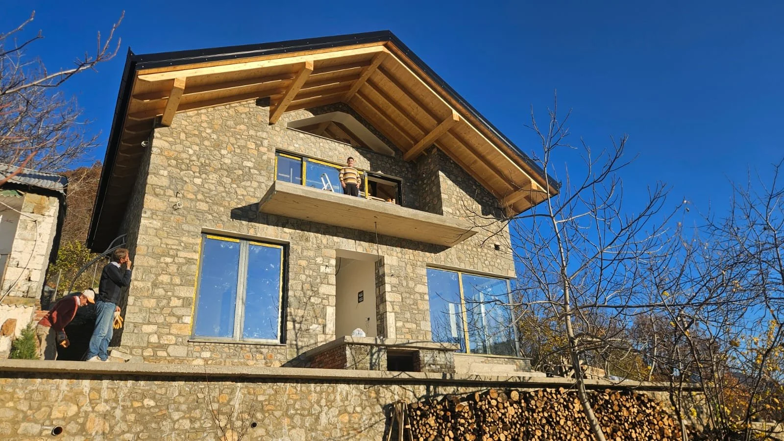 A multi-story house under construction with a stone exterior and large windows, situated on a hillside with leafless trees and a clear blue sky.