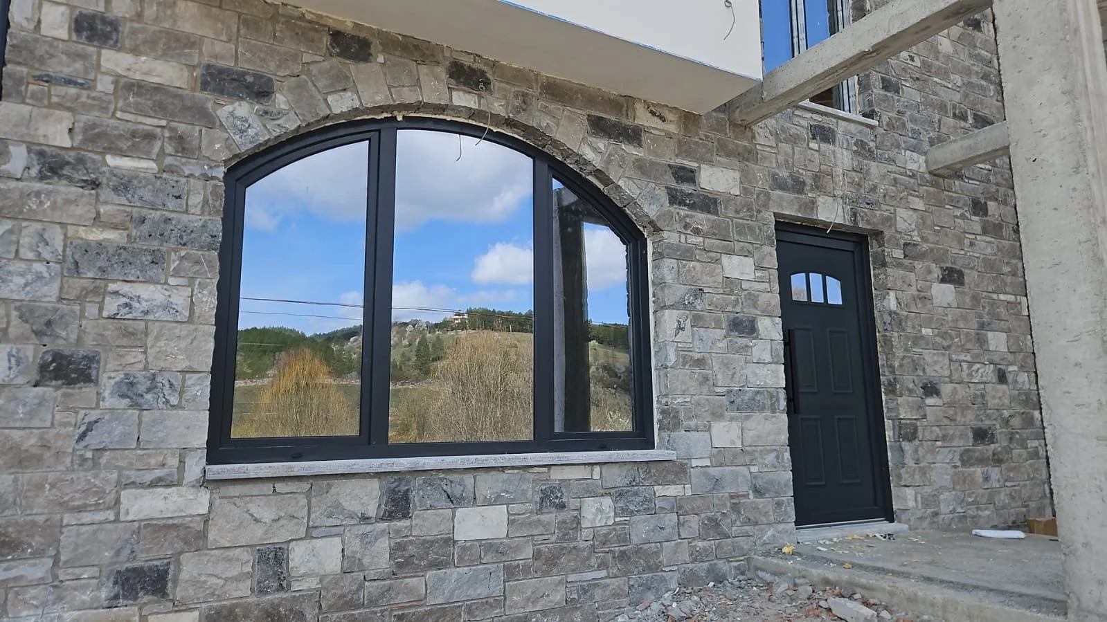 View of a building with a stone facade, featuring a large arched window reflecting the sky and trees, and a black door next to it, with construction materials on the ground.