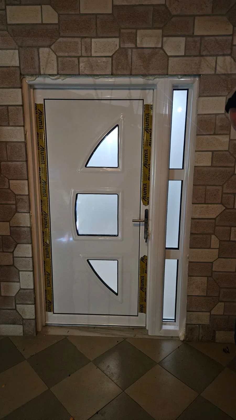 White front door with three small, angled glass windows, surrounded by a brick wall and a glass side panel, during daytime.