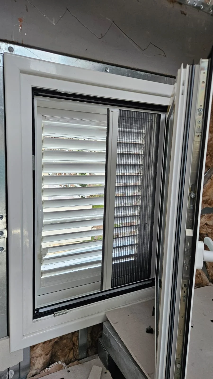 A partially installed black-frame window with white shutters and a sliding insect screen, set in a wall with insulation and construction materials visible around.