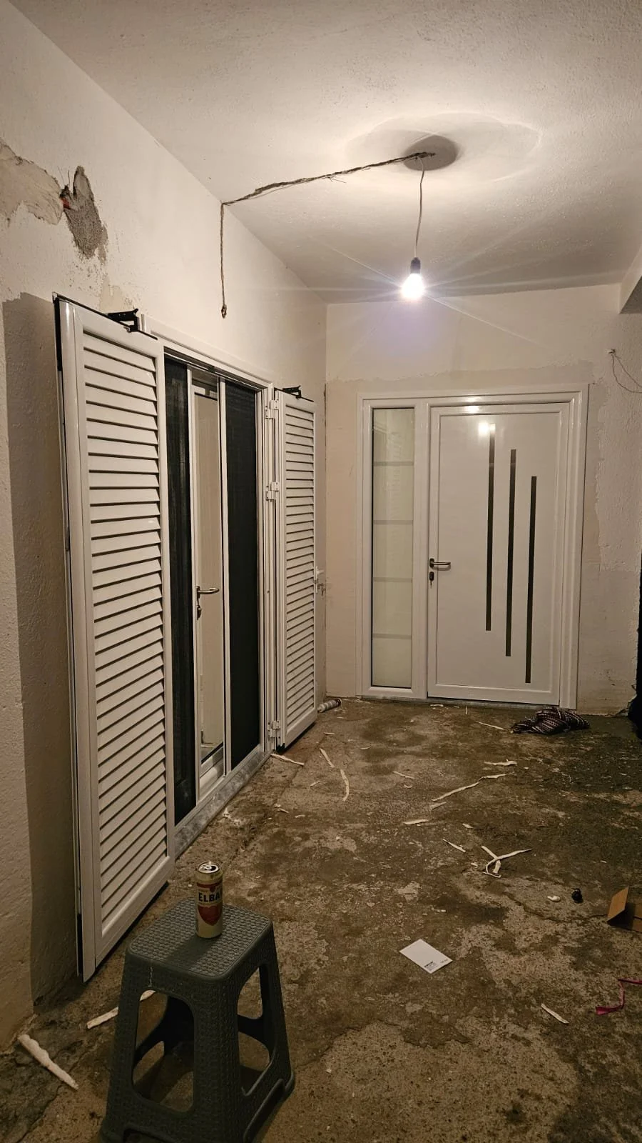 Unfinished room with a single lightbulb hanging from the ceiling, a small stool with a beer can, white double doors with glass panes, and wall patches ready for painting or finishing.