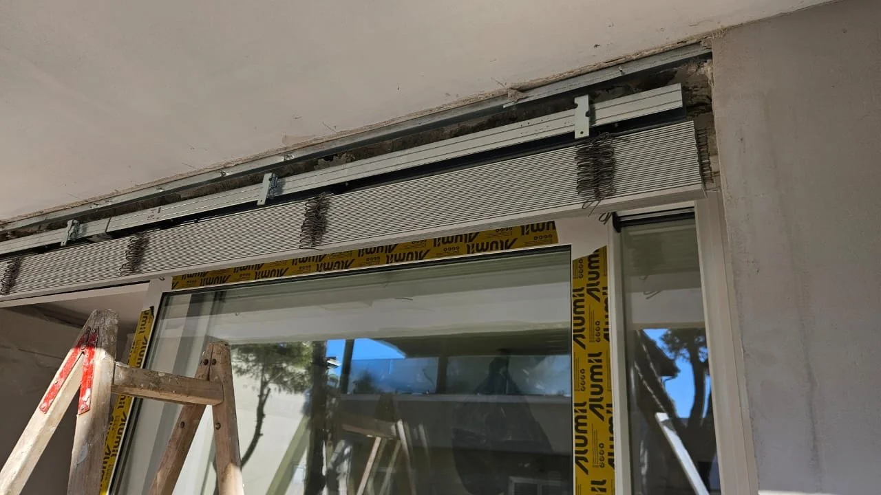 A construction site showing a window installation in progress with an aluminum frame, a wooden ladder, and yellow tape reading 3M, outside visible through the window.