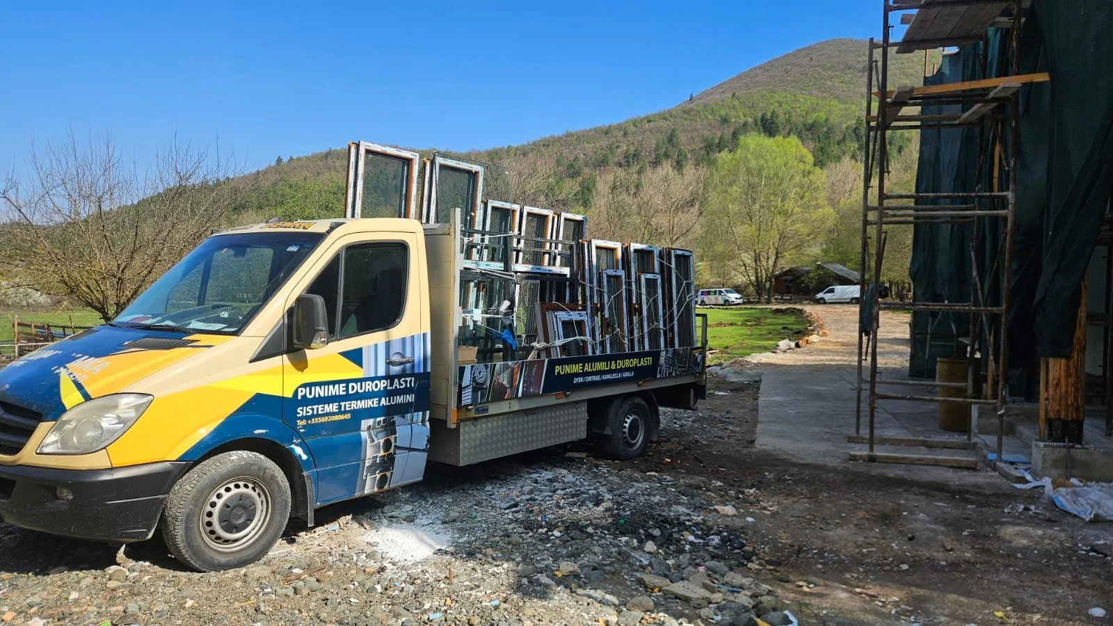 A yellow and blue work truck loaded with multiple window frames parked on a dirt road in front of a construction site with scaffolding, trees, and hills in the background.