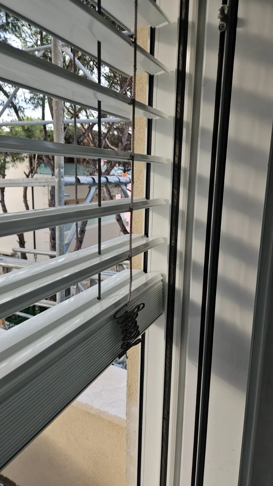 View through window blinds showing construction scaffolding and trees outside, with a portion of a building's exterior wall.
