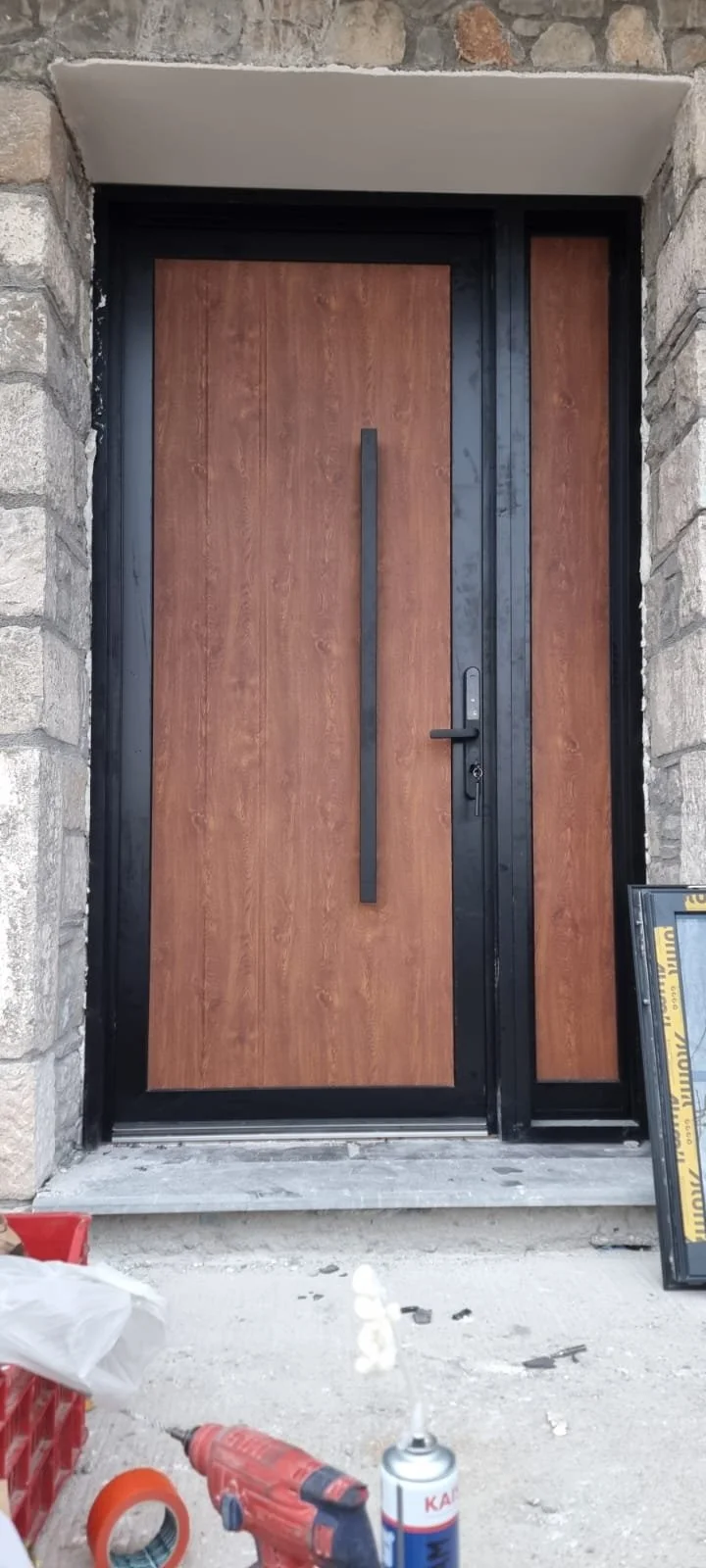 Modern front door with wood finish, black handle, and black frame, set in stone wall, with construction materials in front.