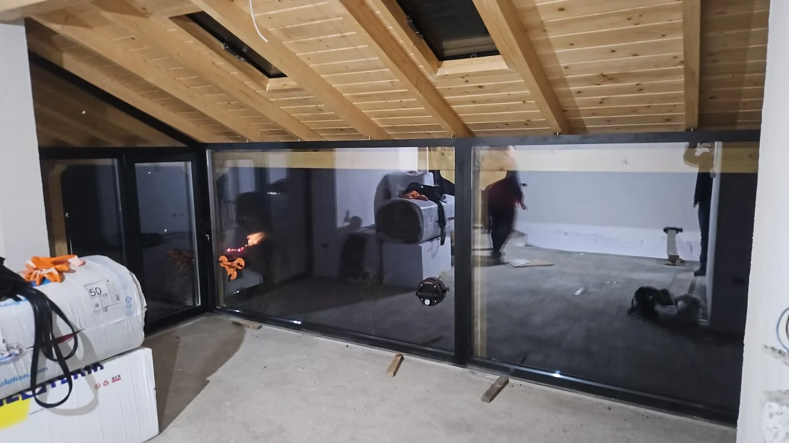 Construction site with glass wall, wooden ceiling, construction materials, and workers installing or inspecting the area.