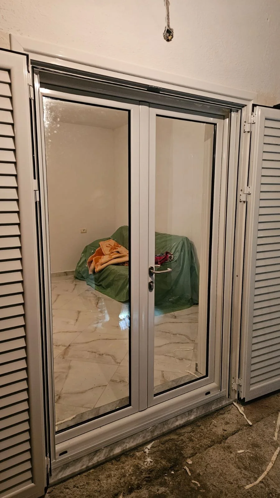 View of an interior room seen through a glass door, with some furniture covered in green plastic and a blanket on top of it.
