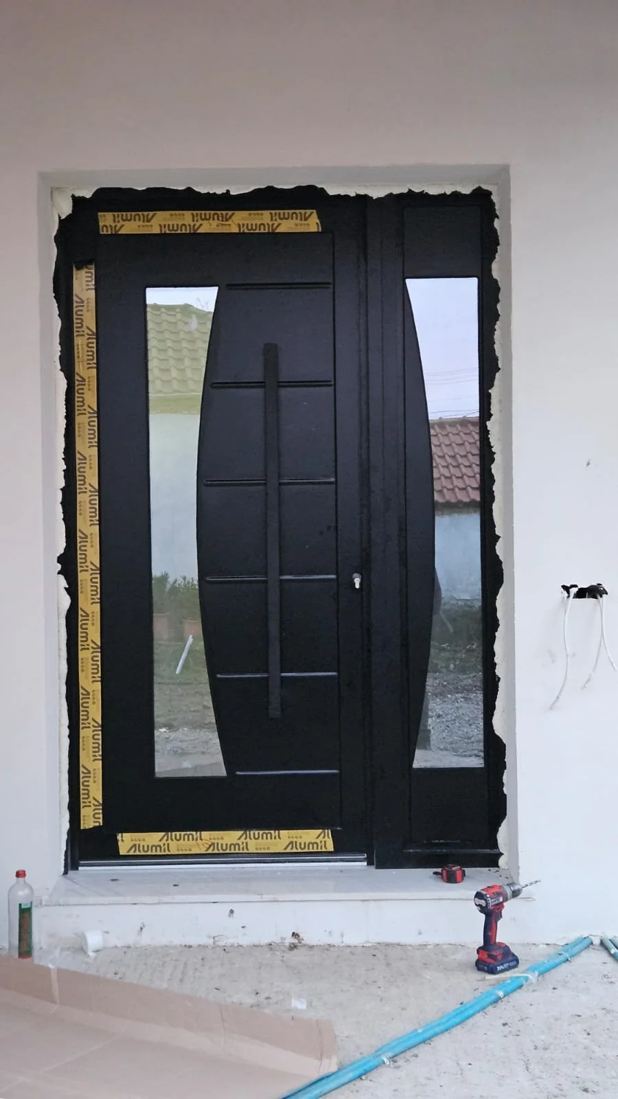 Black front door with glass side panels under construction, with tools and materials nearby, on a white wall with unfinished edges.