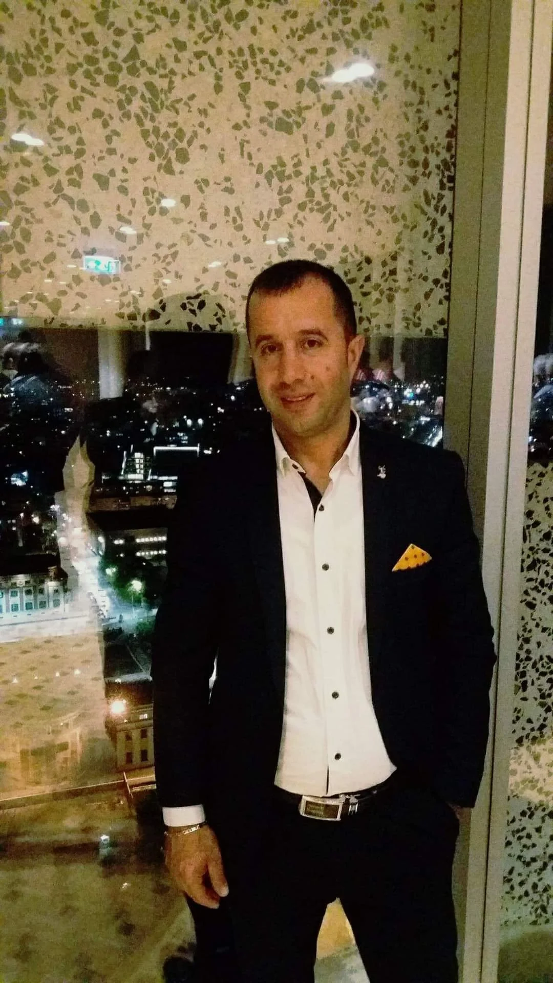 A man in a black suit with a white dress shirt and a yellow pocket square standing indoors during the night, with a cityscape visible through the window behind him. Punime Alumini Demka
