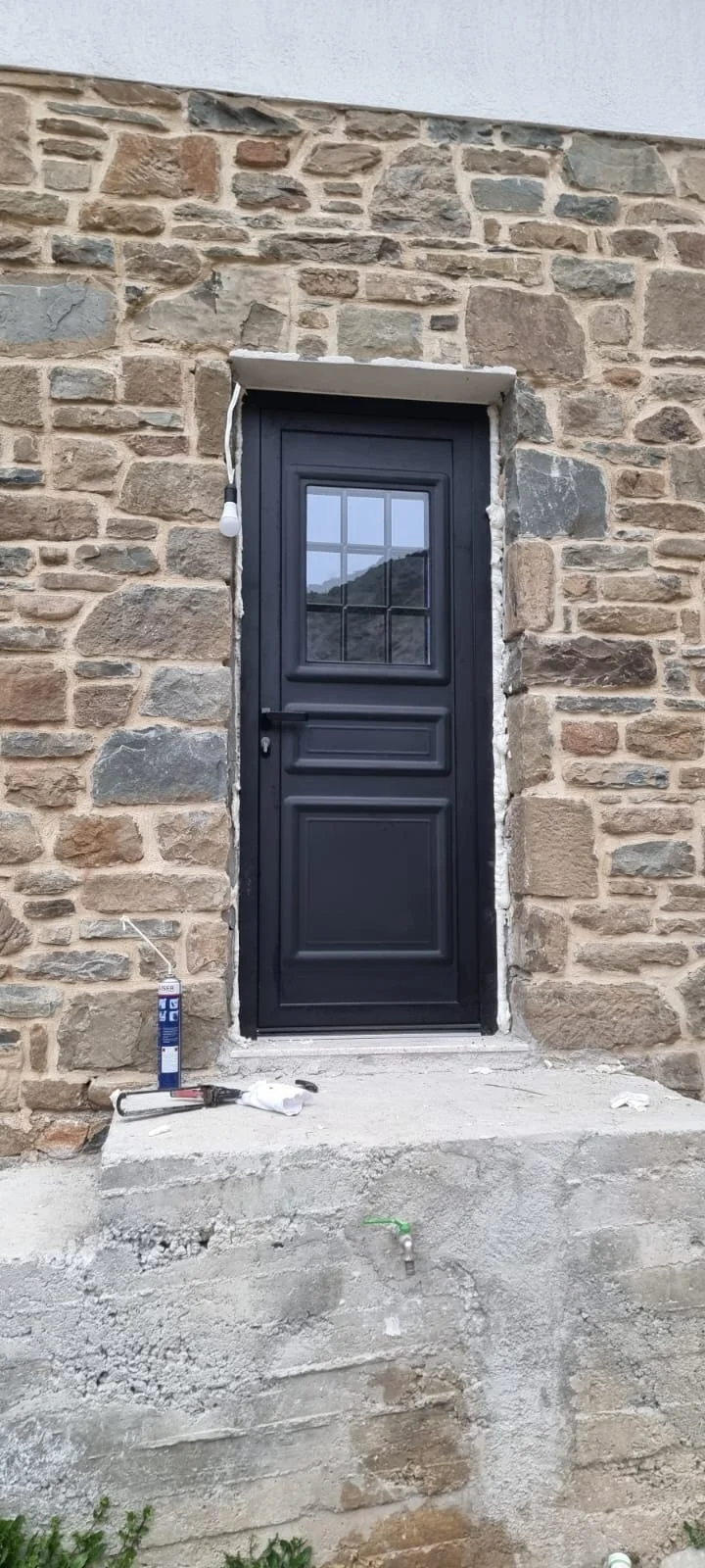 A new black door installed in a stone wall, with construction tools and adhesive caulk on the concrete step in front.