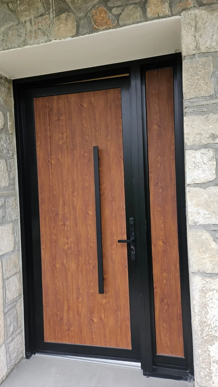 Modern front door with wood finish, black frame, handle, and lock, set in a stone wall exterior.