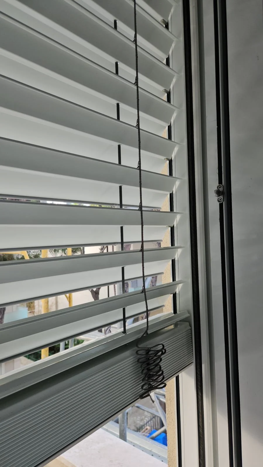 Inside view of a window with white horizontal blinds and a black cord with a spiral-shaped weight hanging from it.