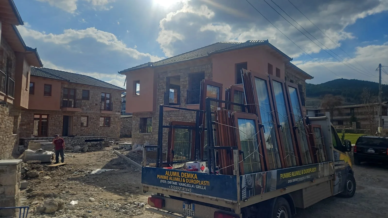 Photo of a construction site with houses, a worker, and a truck carrying windows in a mountainous area under a partly cloudy sky.