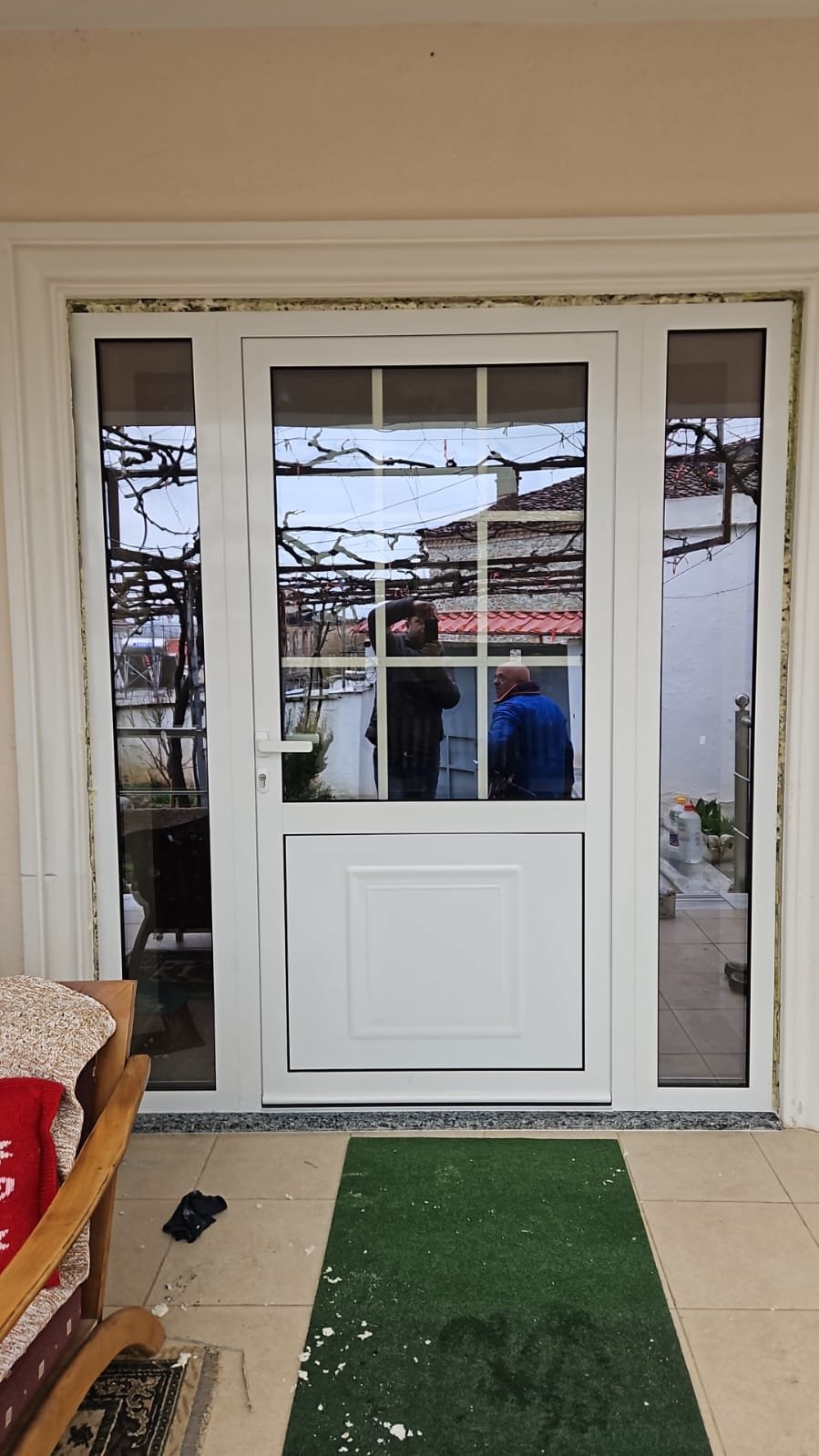 A white patio door with large glass panes reflecting two people, trees, and a house with a red roof outside. Inside, there is a beige tiled floor, a green welcome mat with white splashes, a wooden chair with cushions, and some scattered objects.