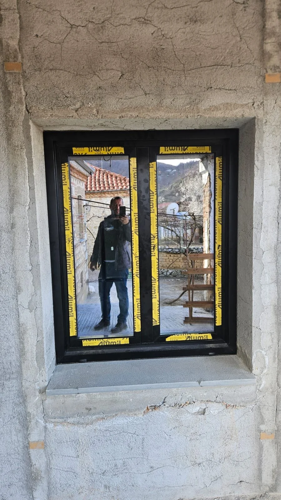 A person taking a photo of their reflection in a newly installed window with yellow protective film, set in a concrete wall with visible cracks.