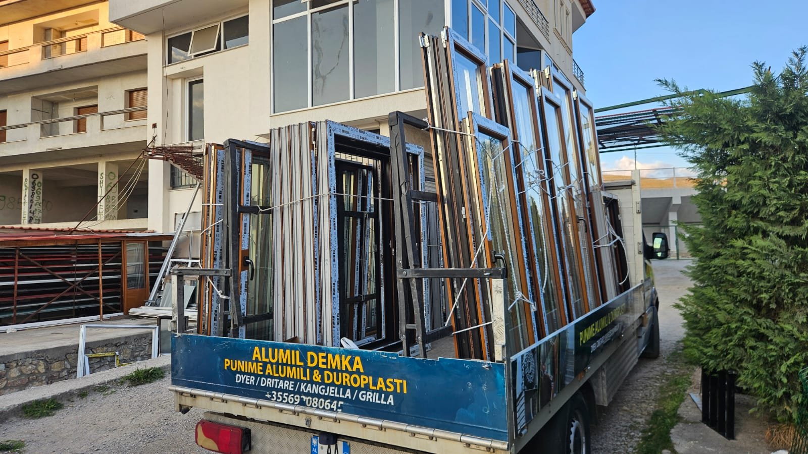 A truck loaded with multiple window frames and glass panes parked outdoors near a modern residential building and a green bush.