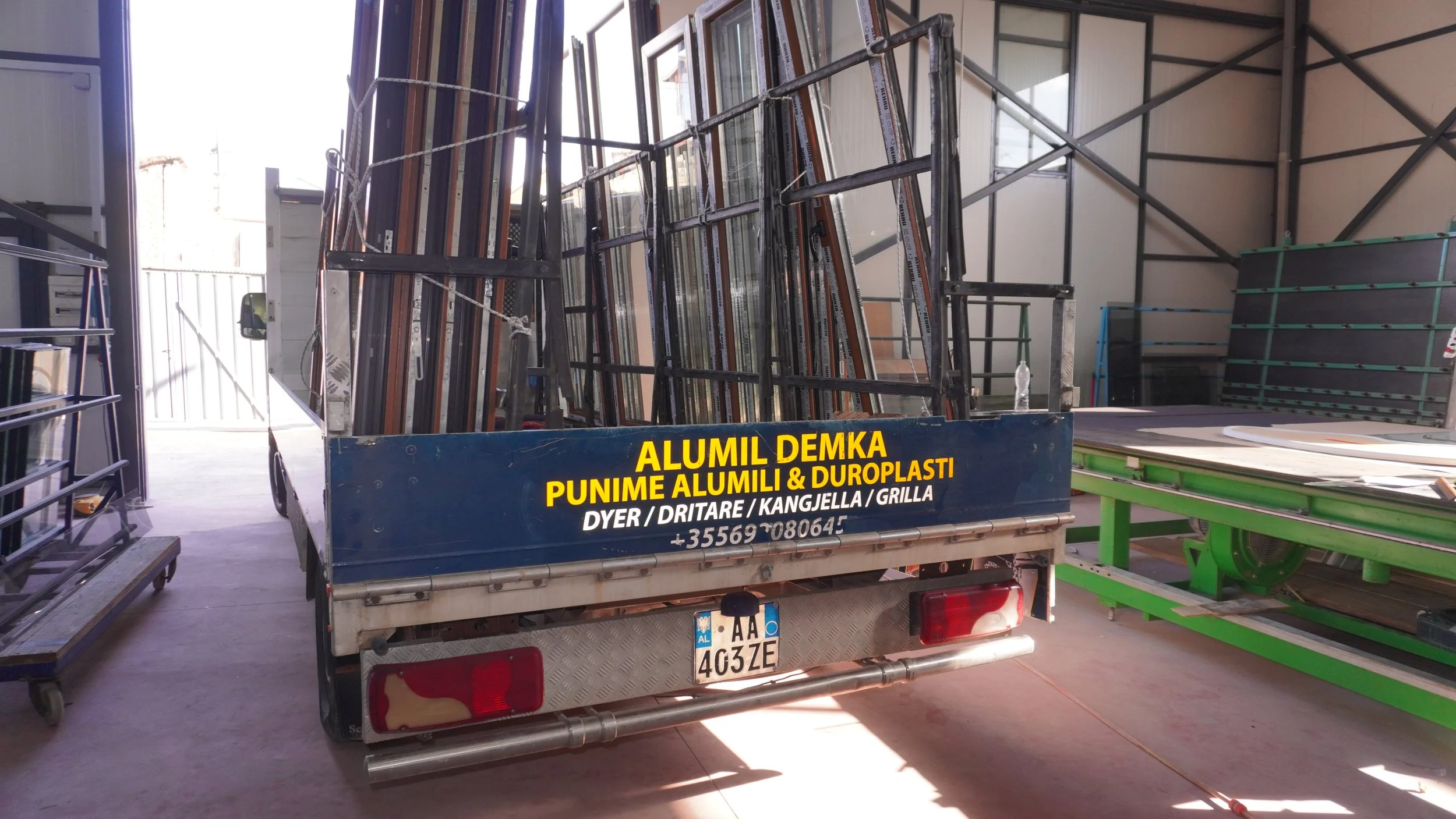 Punime Alumini Demka. A small truck with metal frames and glass in the back, parked inside a warehouse, surrounded by metal and glass products.
