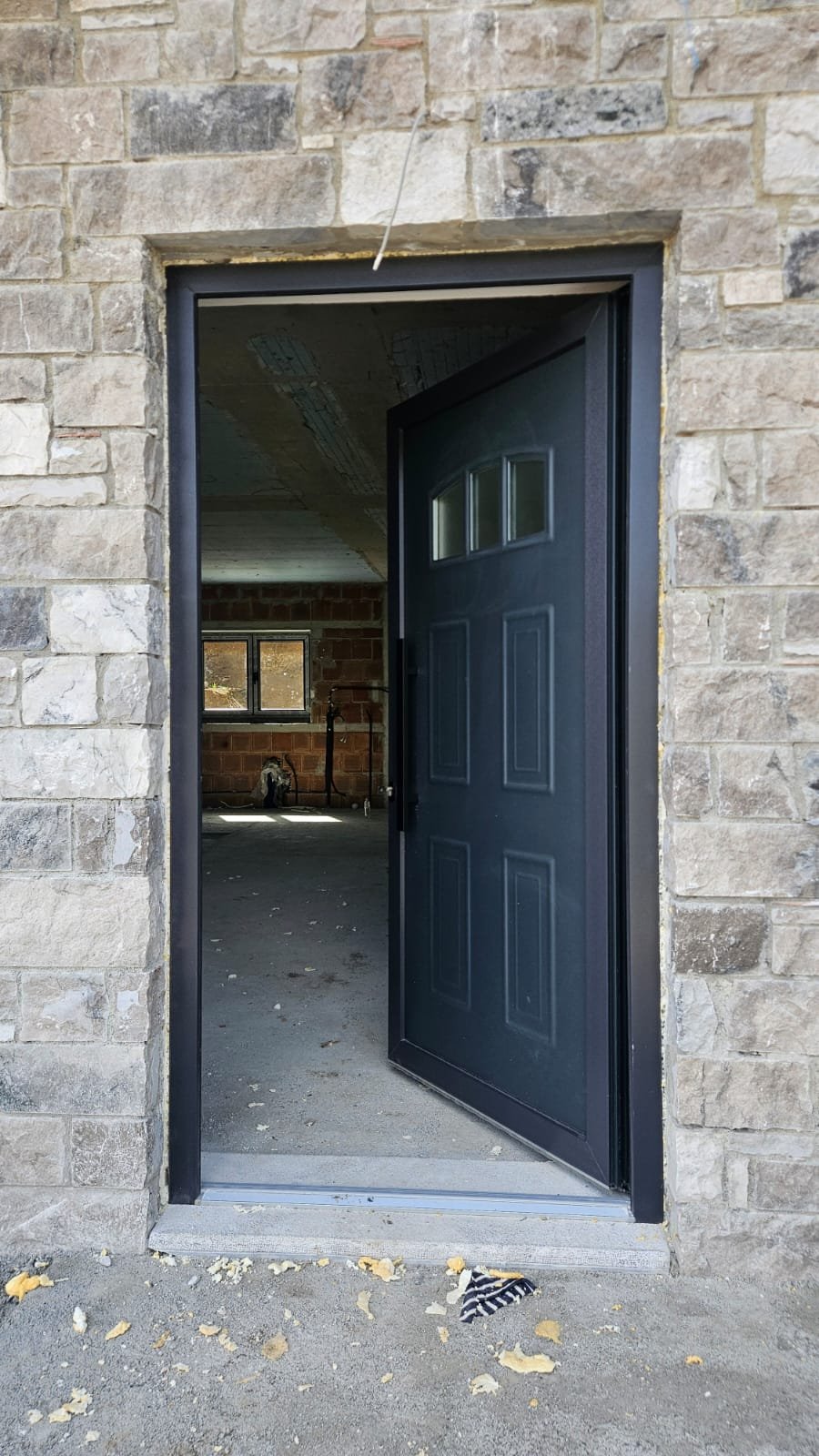 View through a partially open front door into an unfinished interior space with brick walls, a small window, and construction materials on the floor.