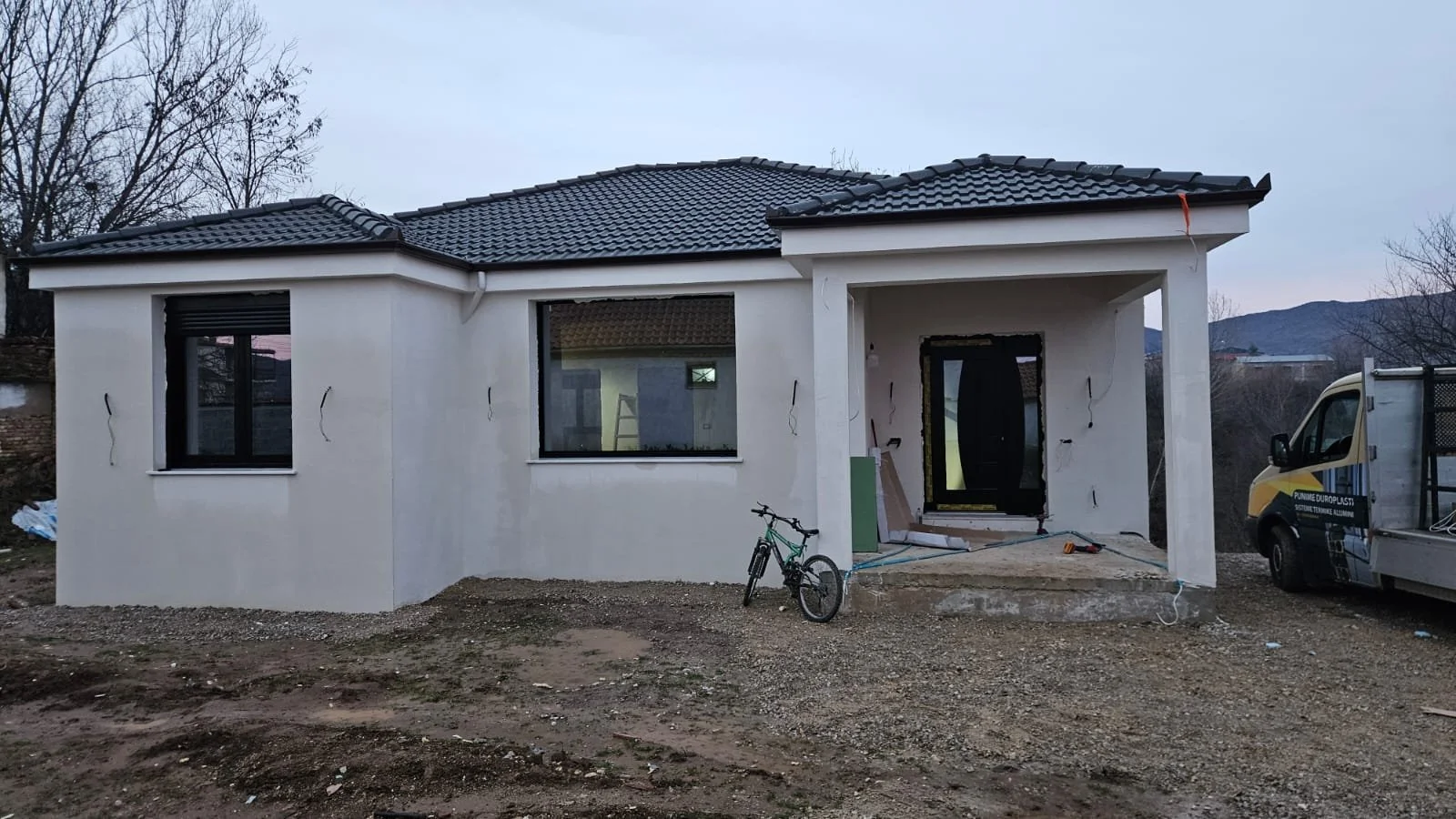 Newly constructed single-family home with white exterior walls, black window frames, and a dark gray tiled roof. The front entrance is still under construction, with visible wiring and unfinished steps. A bicycle is parked in front, and construction 