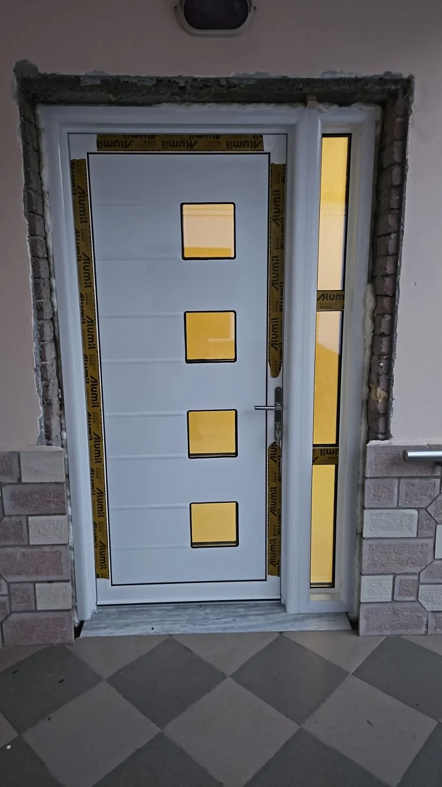 New white exterior door with four square windows, construction stickers around the frame, partially surrounded by brick and concrete walls, and a tiled floor in front.