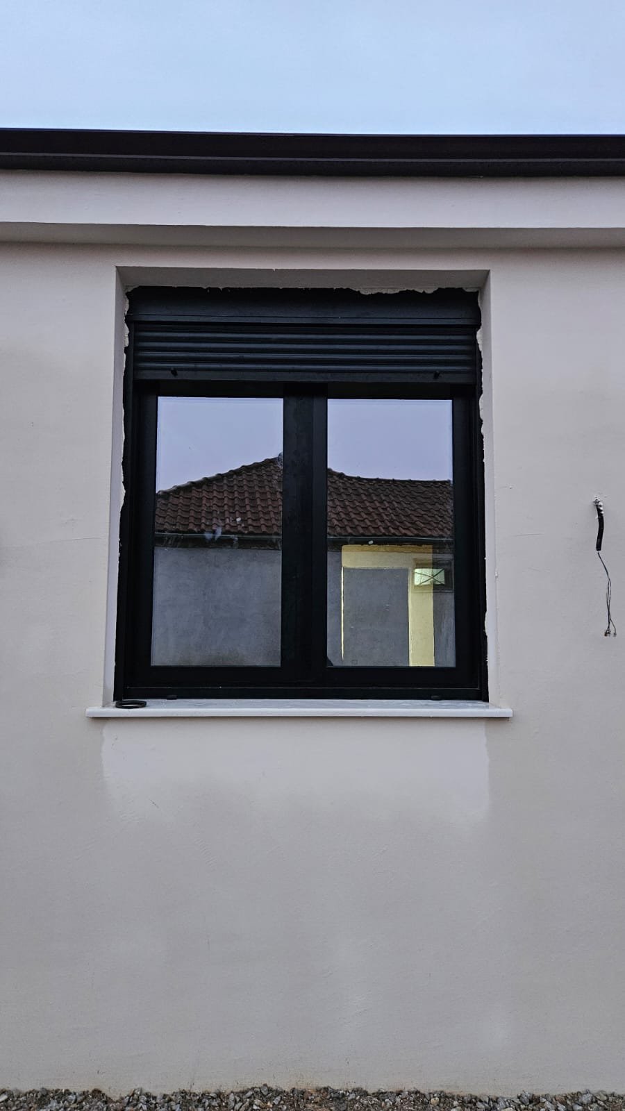 A window in a white wall with black frame and partially closed black shutter, reflecting a neighboring house with a tiled roof.