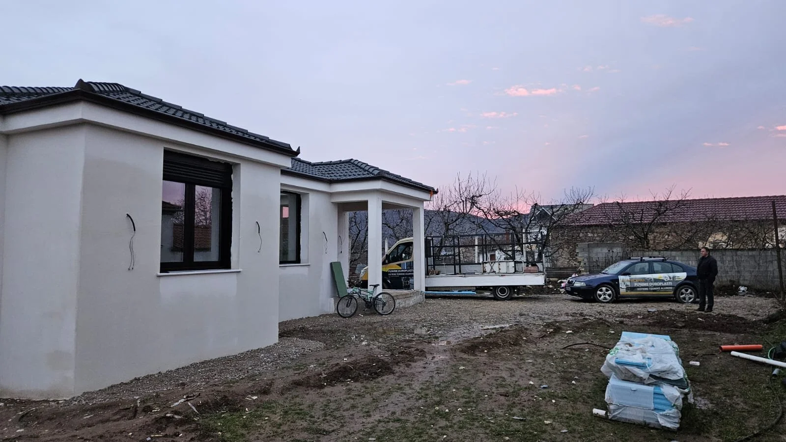 Construction site of a house with white exterior walls, a black tiled roof, and multiple windows. There is construction material on the ground, a bicycle, a truck with a flatbed, and a person standing near a car with a yellow and black sign. The sky 