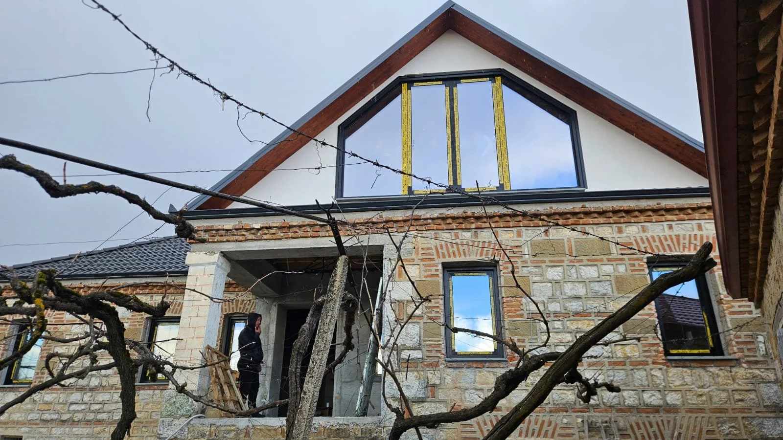 A house under construction with a large front window, brick and stone exterior, and a person standing near the entrance. There are bare tree branches in the foreground.