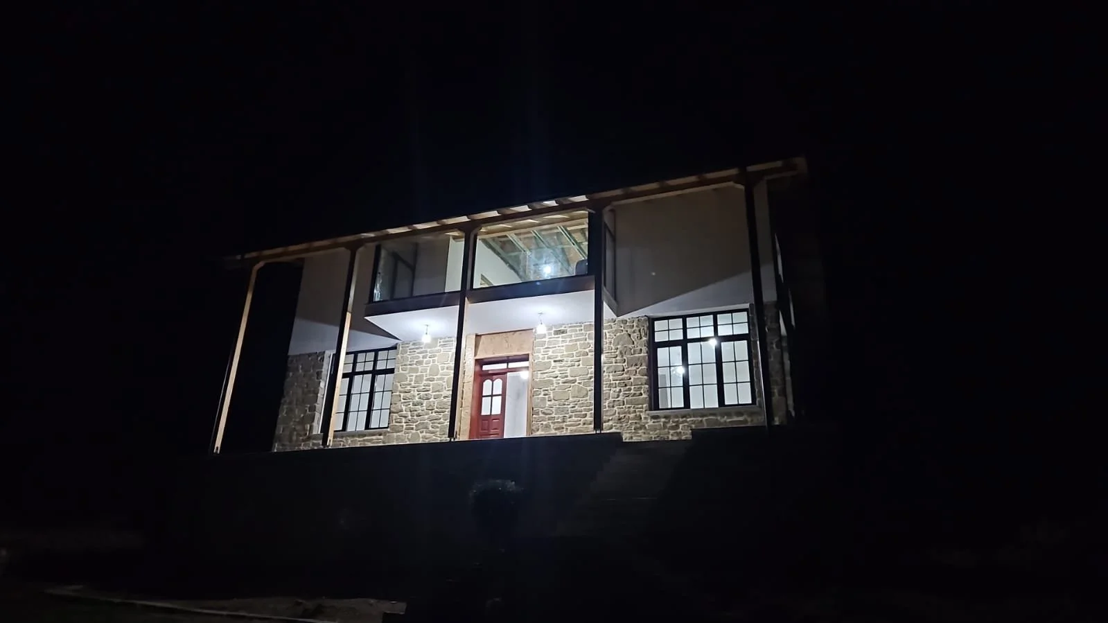 A house under construction at night with lights on, stone walls, large windows, and a red front door, elevated on a platform with stairs.