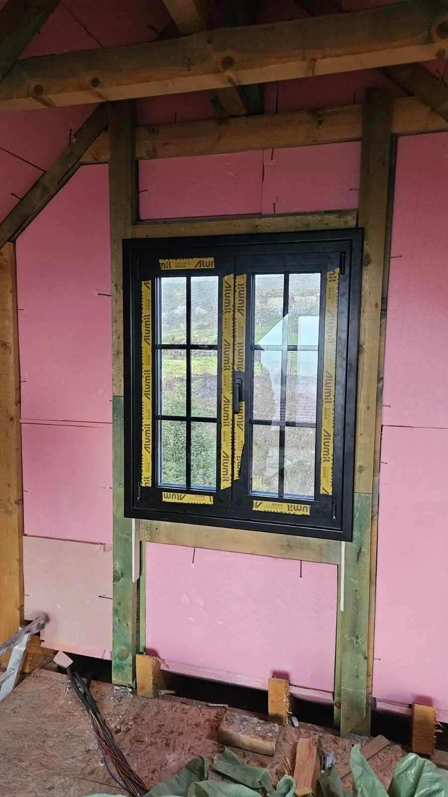 A black window installed in a wooden frame inside a building under construction, with pink insulation on the walls and construction materials and wiring on the floor.